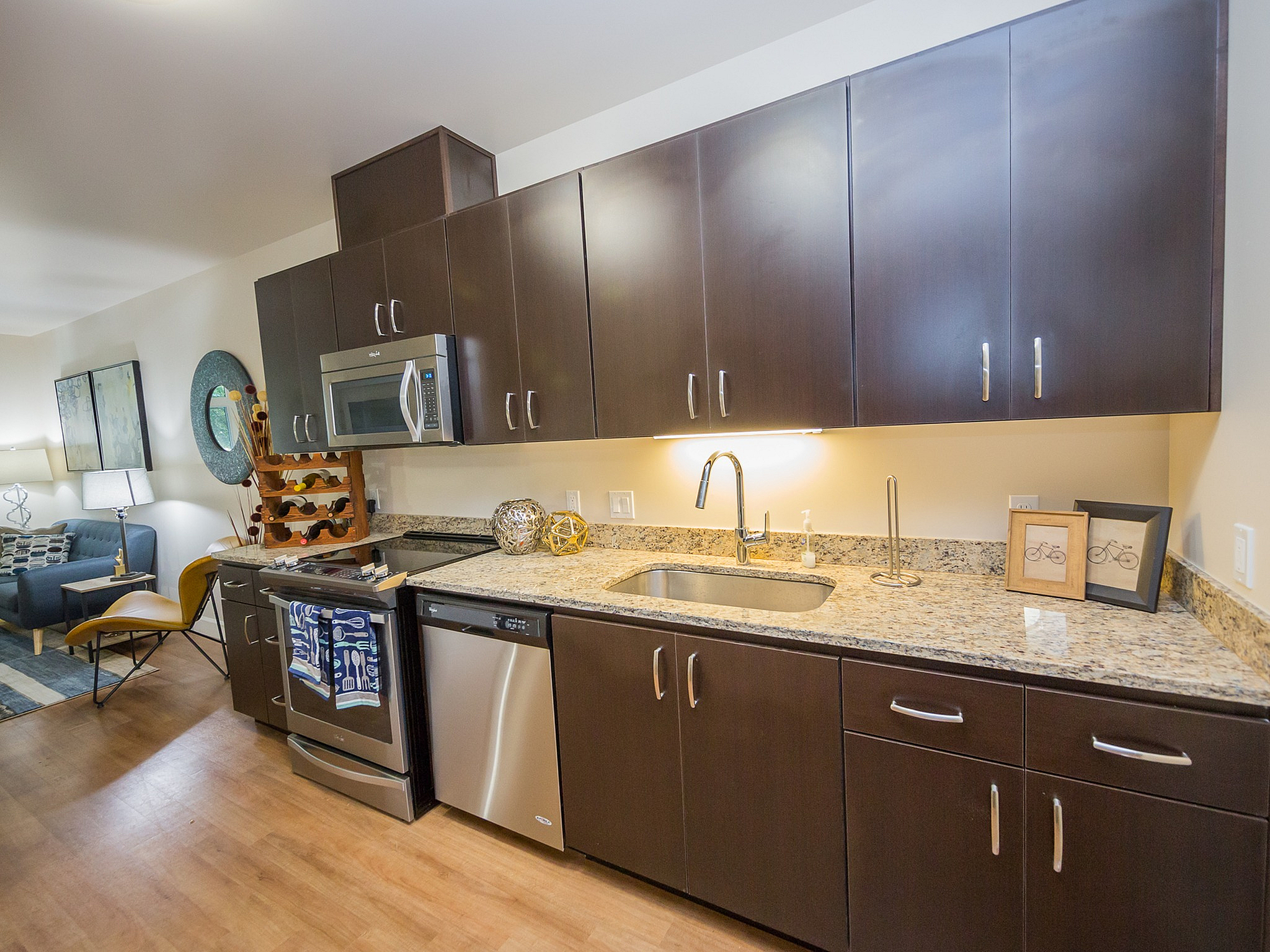 Modern kitchen with sleek dark wood cabinets, stainless steel appliances, and spacious granite countertops, opening into a cozy living area.