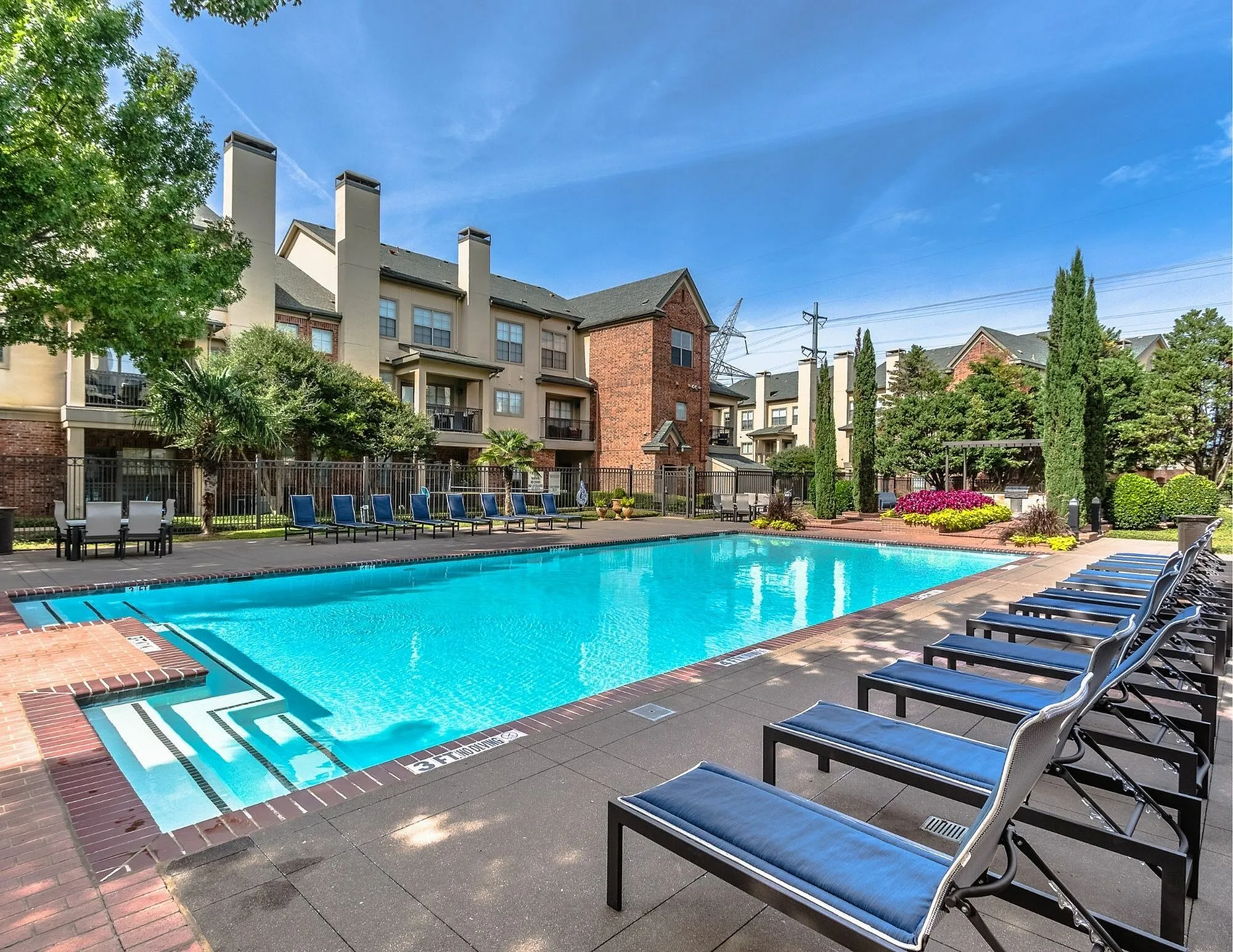 Luxurious apartment complex featuring a sparkling pool with sun loungers, surrounded by lush greenery and elegant architecture.