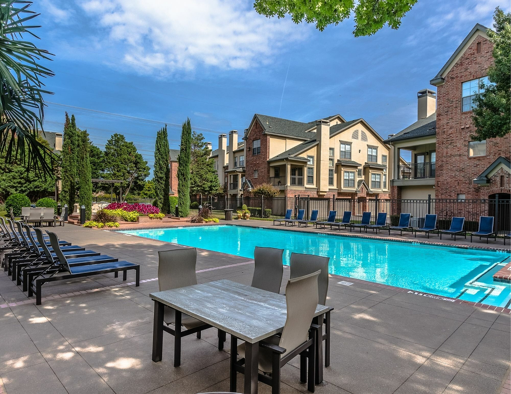 Luxurious apartment community with a stunning outdoor pool, surrounded by elegant lounge chairs, lush landscaping, and charming brick architecture.