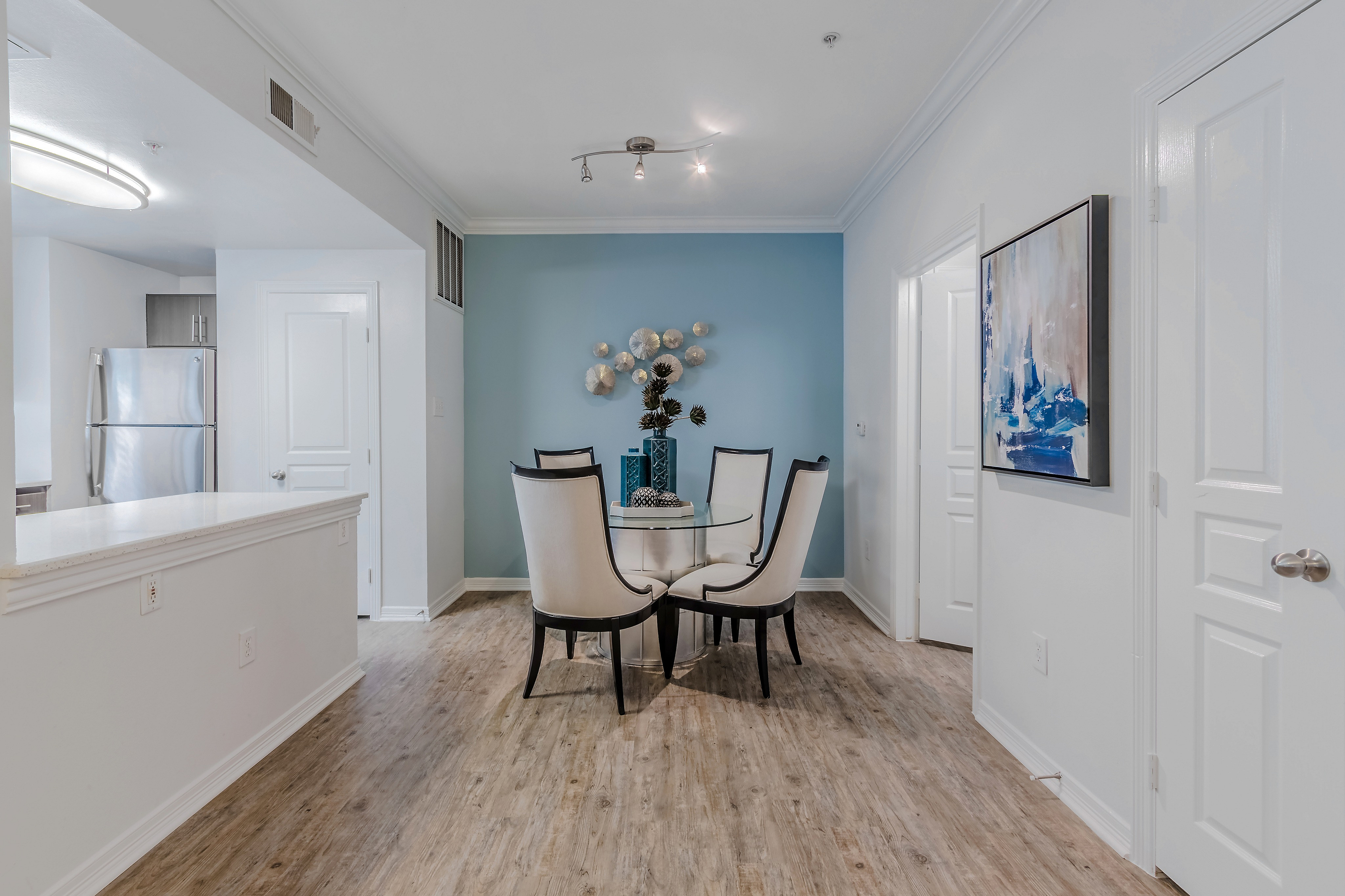 Chic dining area with modern decor, featuring a glass table and elegant chairs, set against stylish blue accent wall in a spacious apartment.