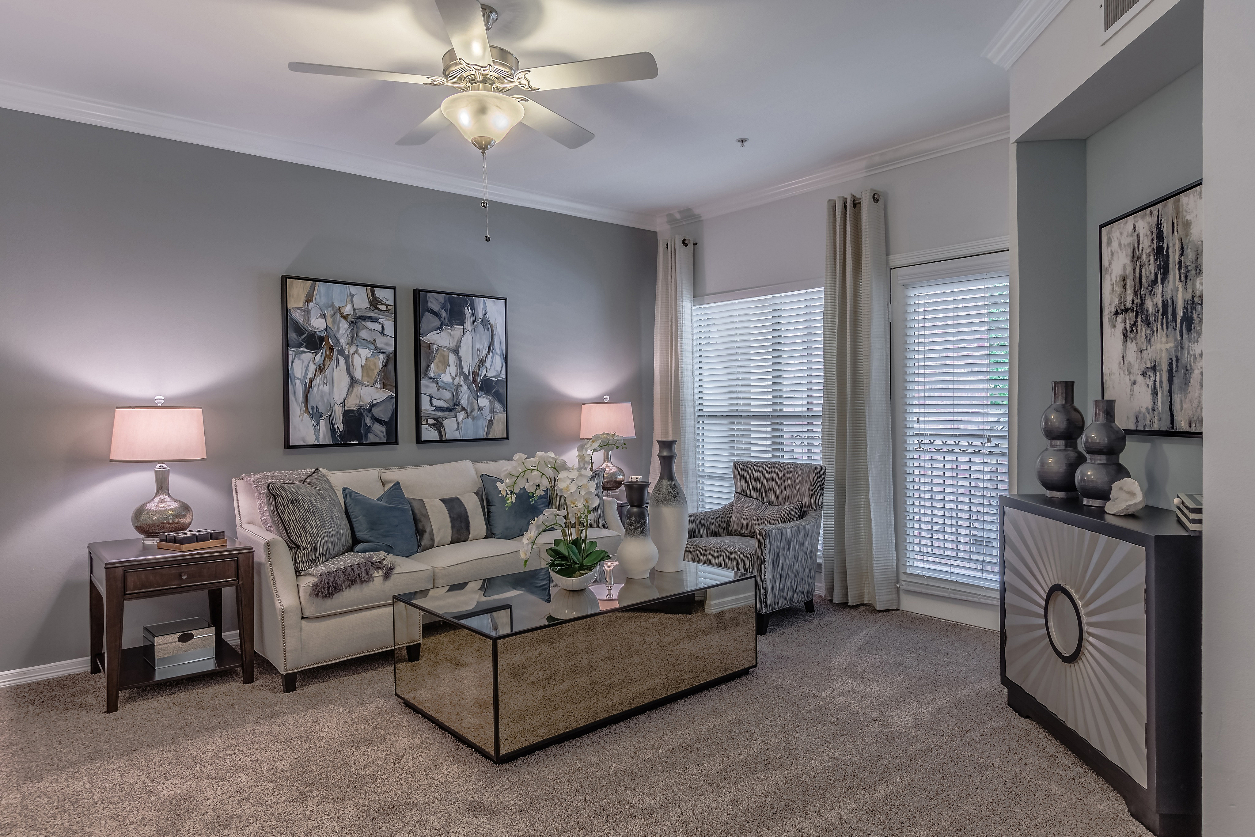 Elegant living room with plush beige sofa, abstract artwork, and stylish glass coffee table under a ceiling fan.