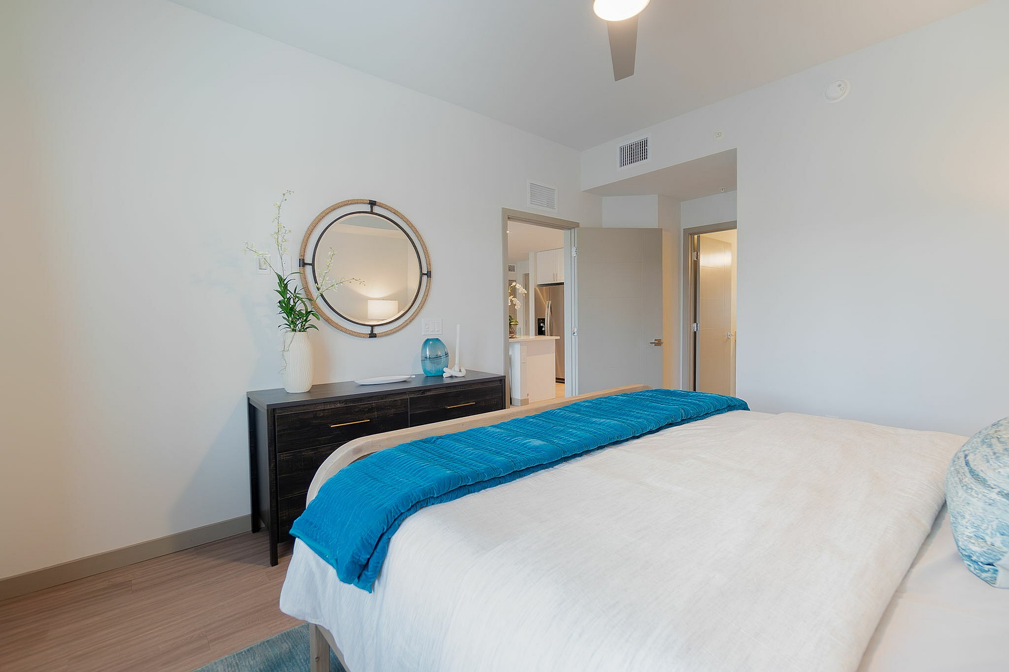 Inviting bedroom with plush bedding, a stylish mirror, and a sleek dresser, offering a glimpse into a modern open-concept kitchen.