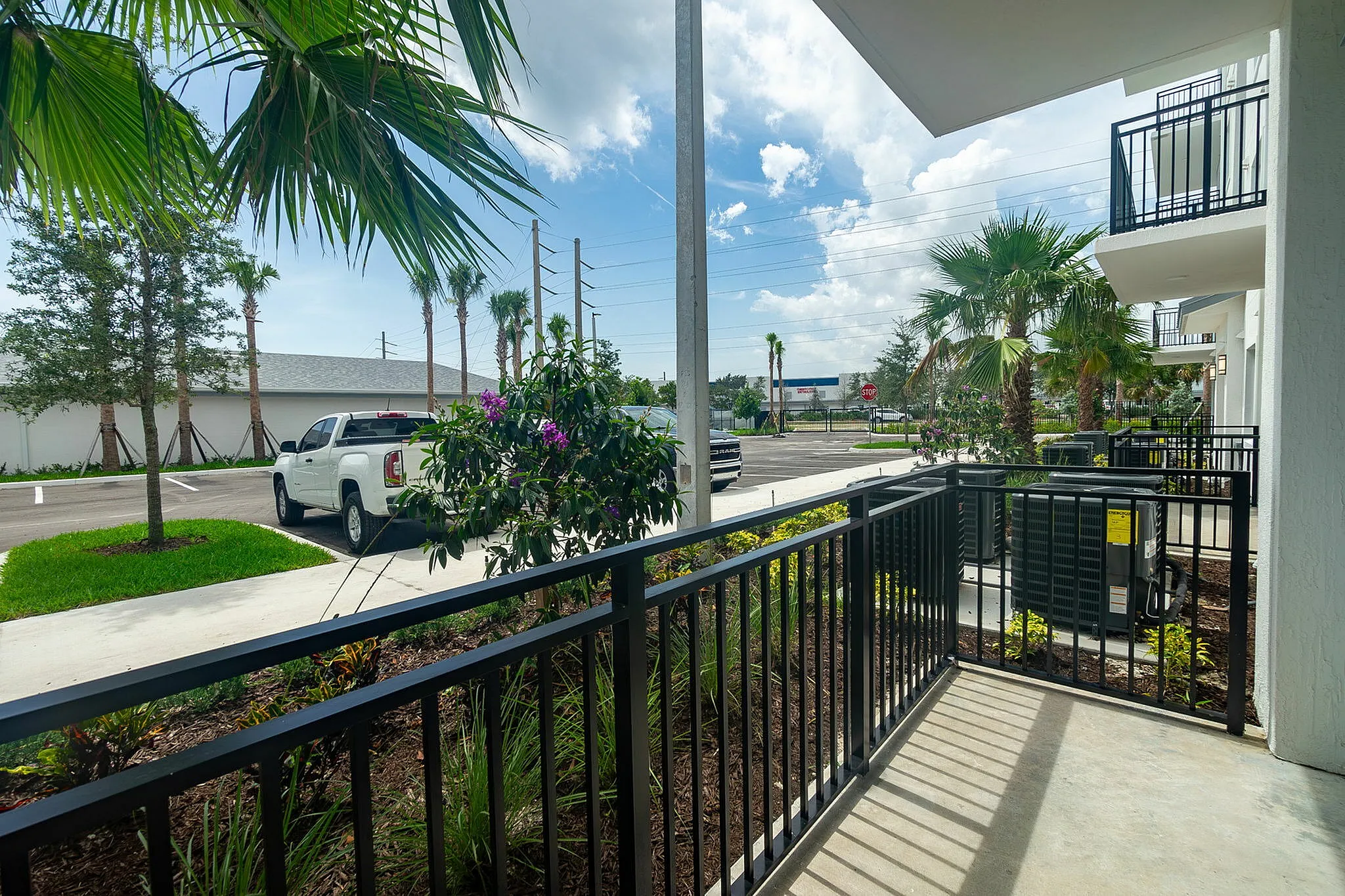 Charming apartment balcony overlooking lush landscaping and a serene suburban street, featuring vibrant plants and palm trees under a bright blue sky.