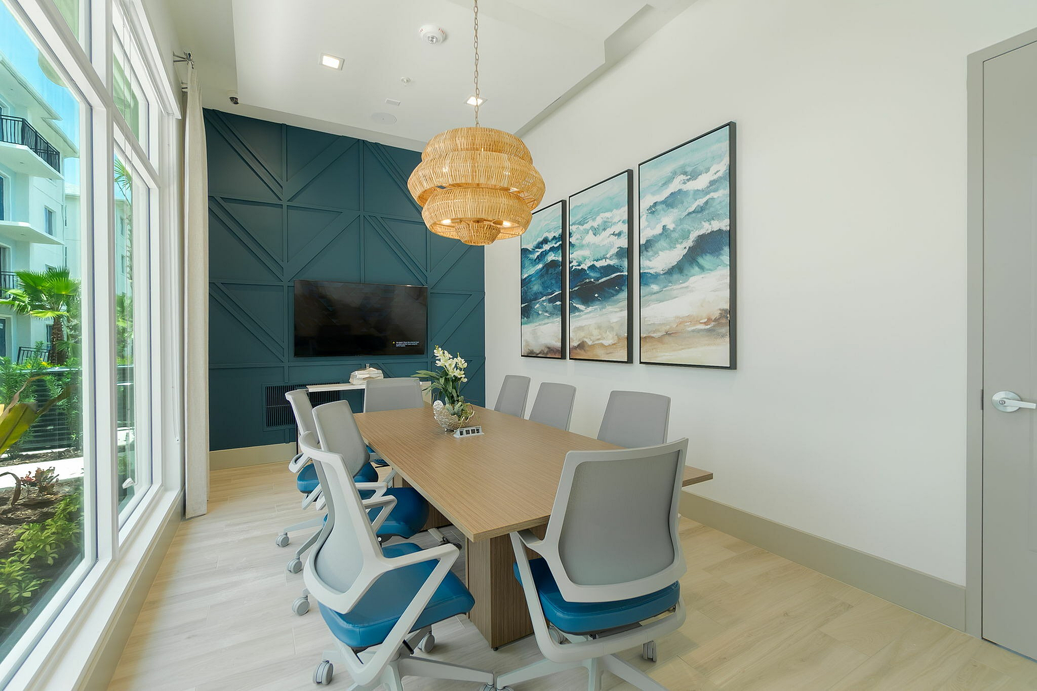 Modern conference room featuring elegant wooden table, stylish rolling chairs, large windows with outdoor view, decorative ocean-themed art, and contemporary pendant lighting.