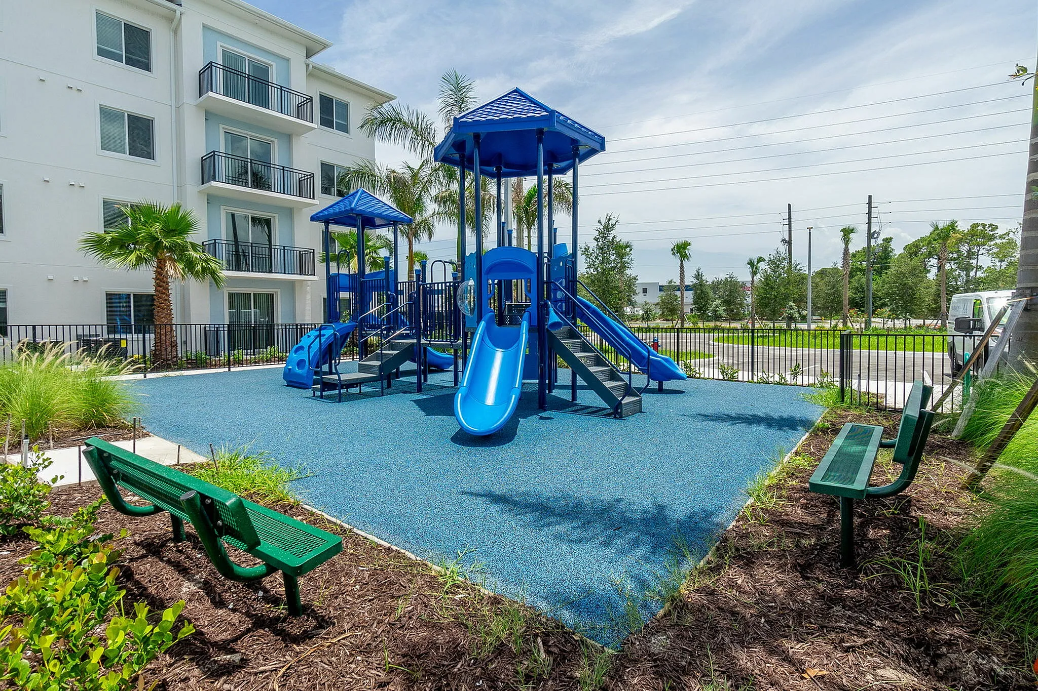 Modern apartment complex with a vibrant blue playground, surrounded by lush greenery and palm trees, perfect for family living.