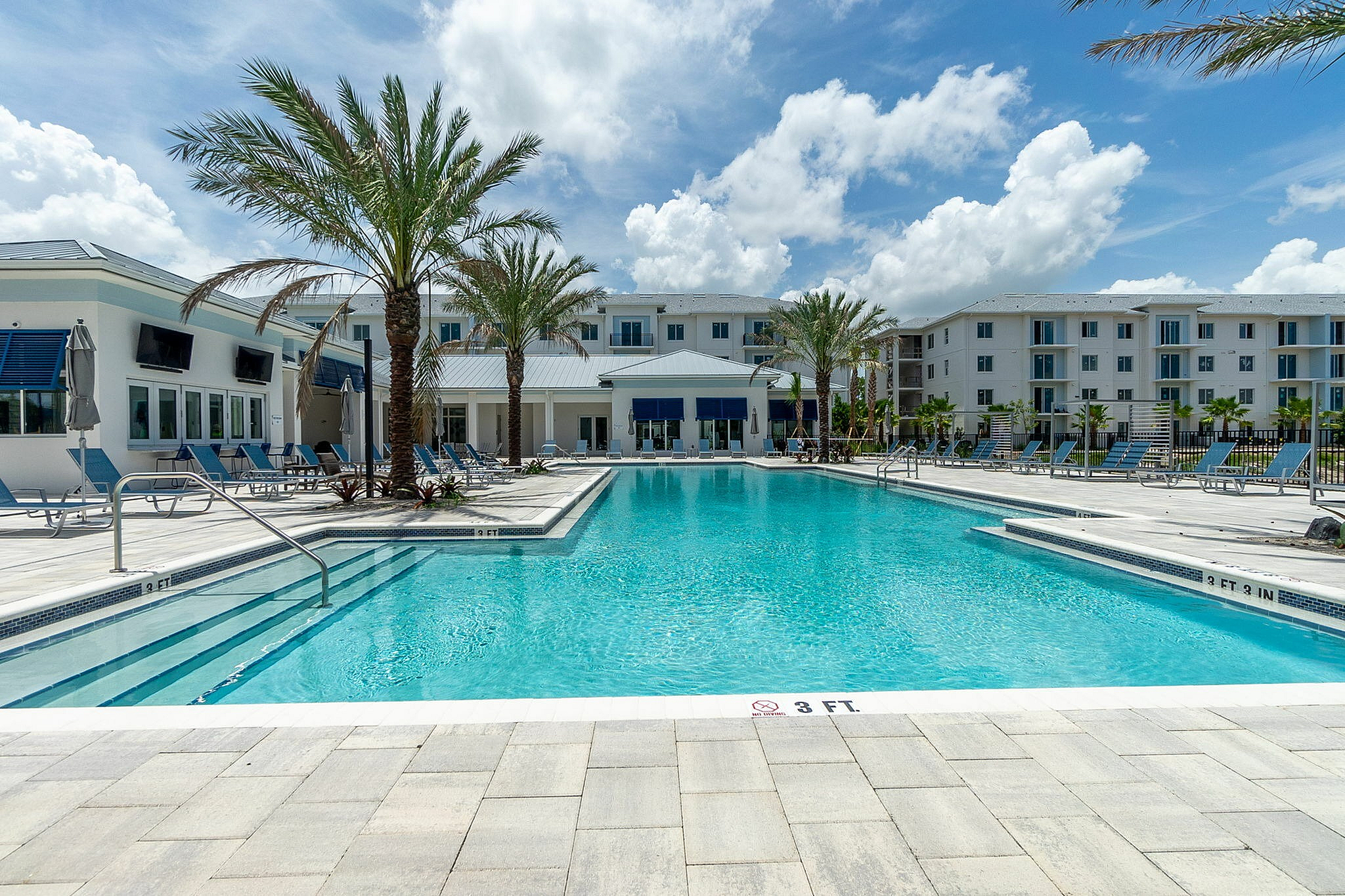 Luxurious apartment complex with a stunning resort-style pool, elegant poolside loungers, and beautiful palm trees under a bright blue sky.