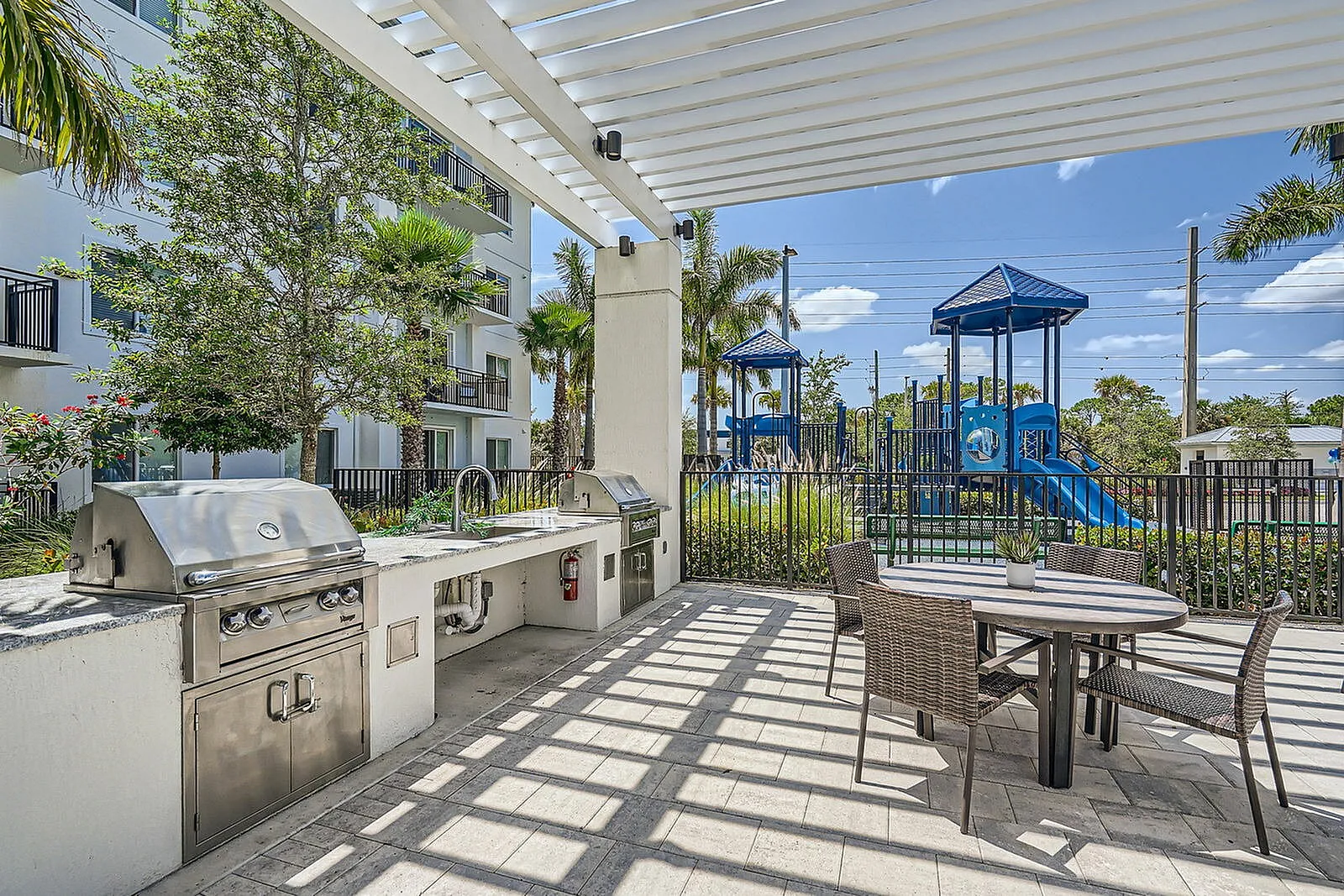 Modern outdoor kitchen and dining area with stainless steel grills and seating under a stylish pergola, next to a vibrant playground surrounded by lush greenery.