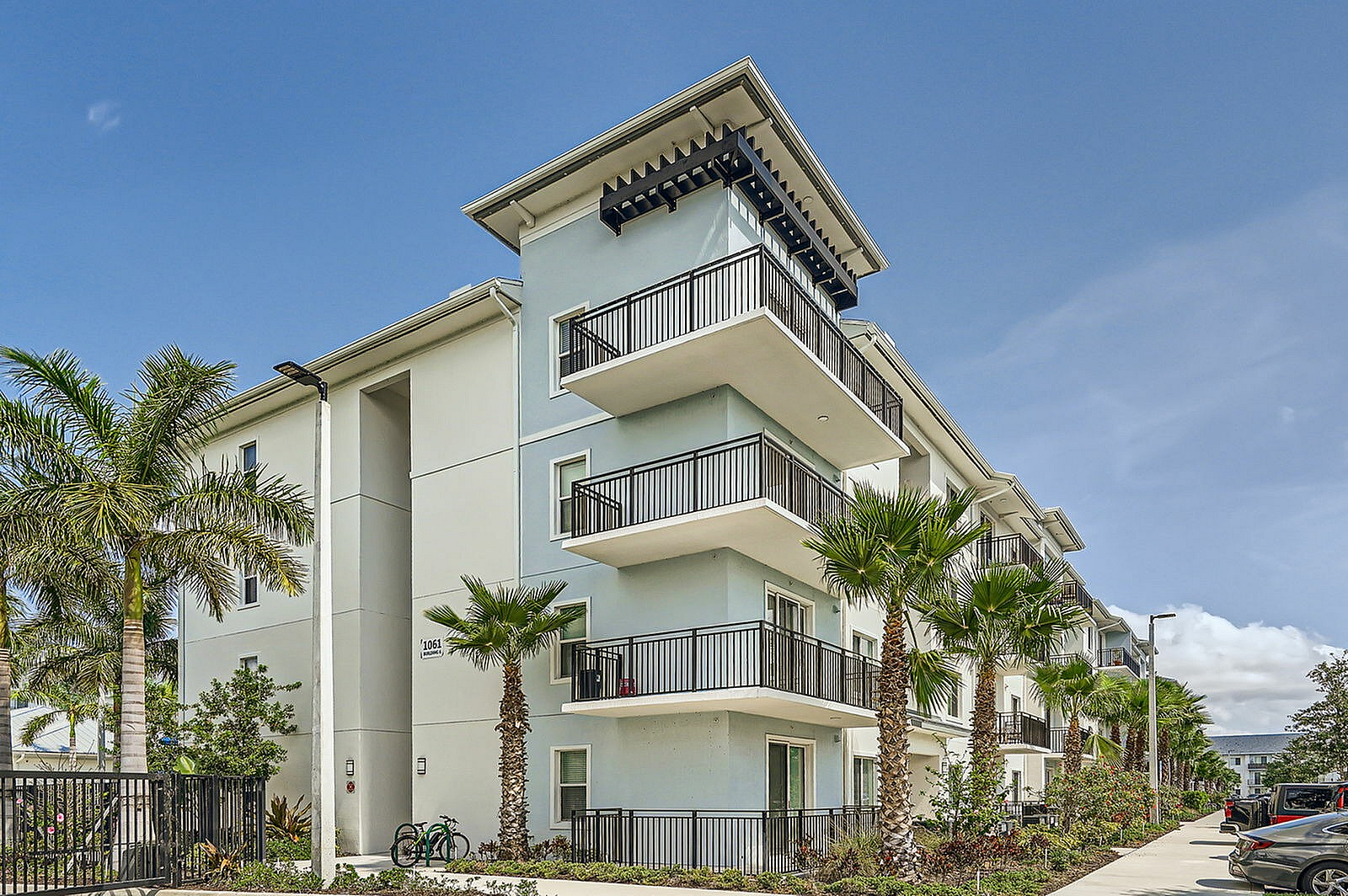 Modern apartment building with spacious balconies, surrounded by lush palm trees under a bright blue sky.