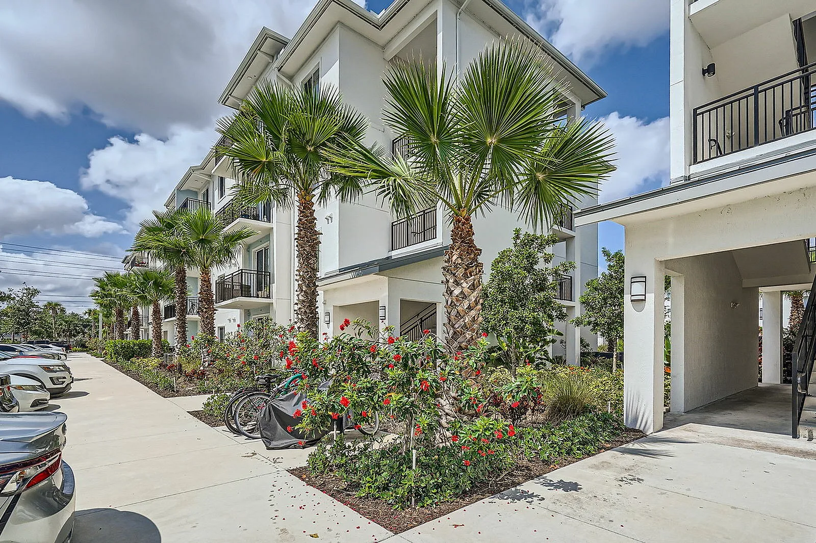 Modern apartment building with lush palm trees and vibrant flowers, featuring sleek balconies and reserved parking in a sunny location.