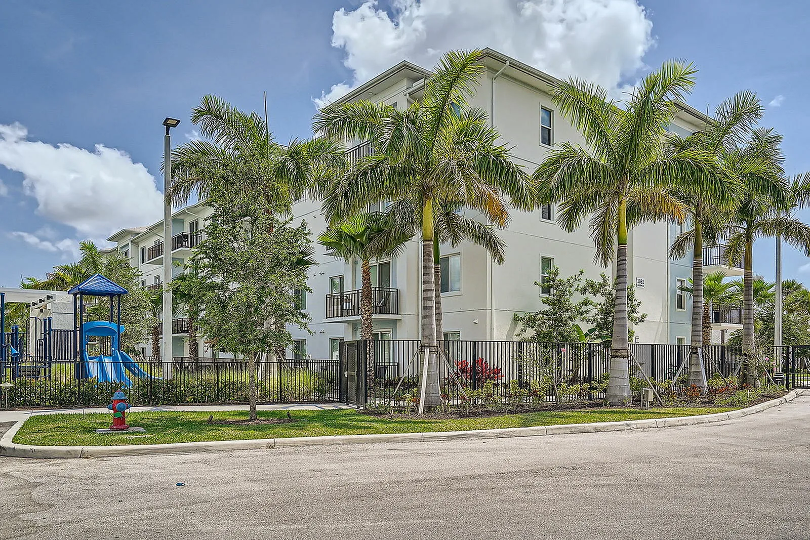 Bright and modern apartment building surrounded by lush palm trees and a colorful playground, offering a perfect blend of tranquility and family-friendly amenities.