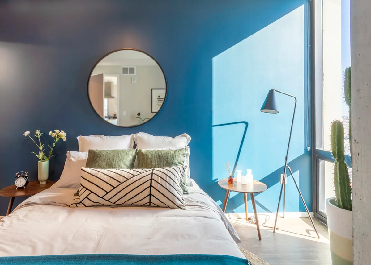 Bright and inviting bedroom with modern decor, featuring a cozy bed, stylish round mirror, and abundant natural light through large window.