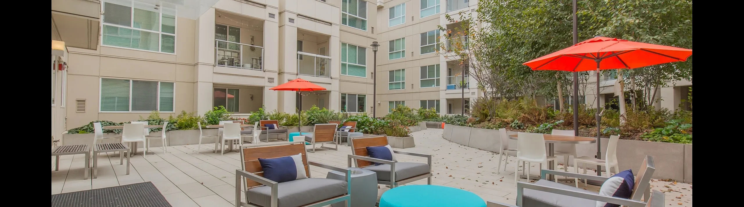 Modern apartment courtyard with stylish outdoor seating, vibrant red umbrellas, and lush greenery, perfect for relaxation and social gatherings.