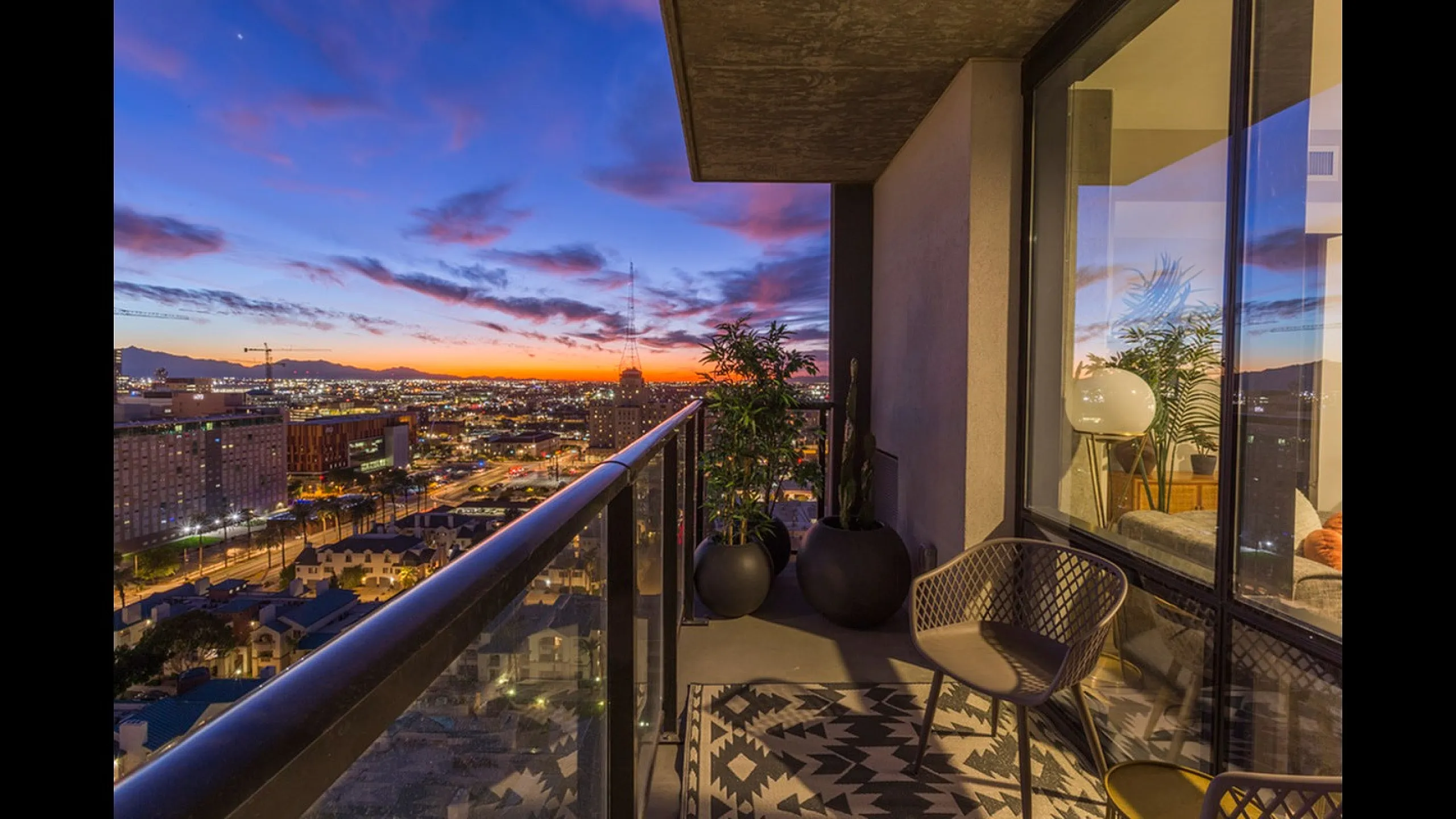 Stunning cityscape view from a modern apartment balcony at twilight, featuring elegant outdoor seating and lush potted plants.