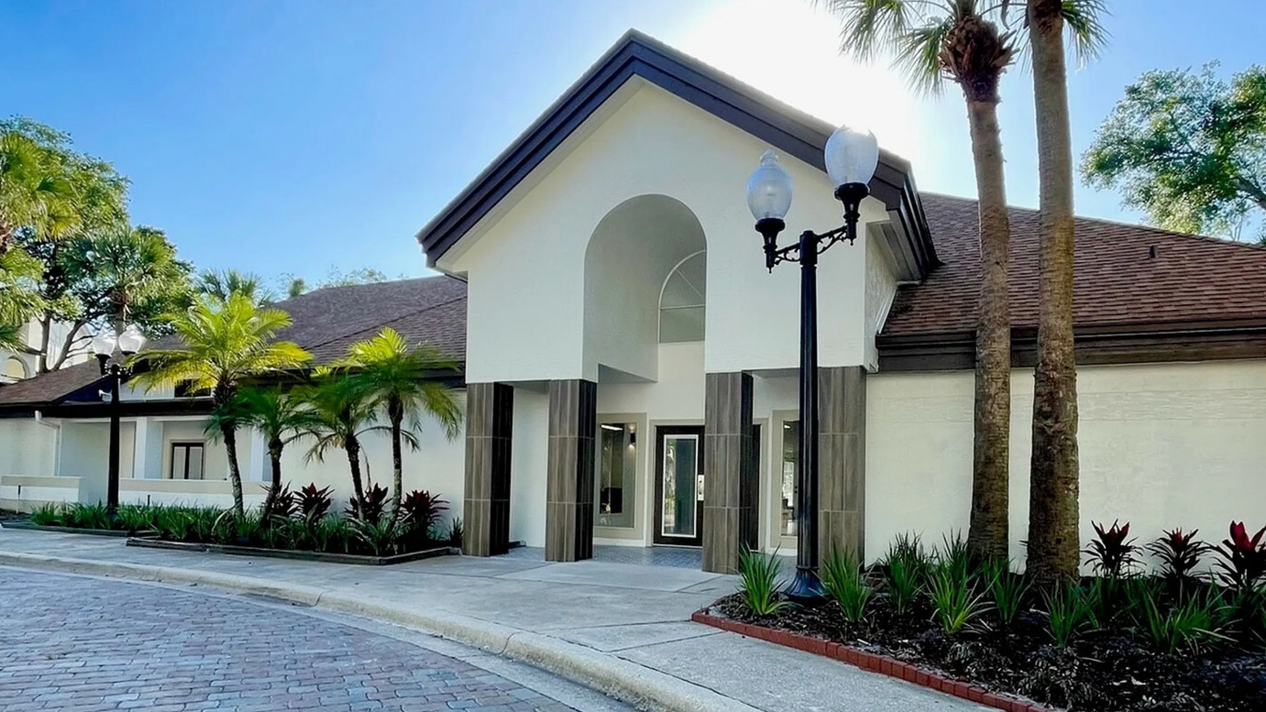 Elegant entrance to a luxurious apartment complex, featuring palm trees and a charming cobblestone driveway under a clear blue sky.