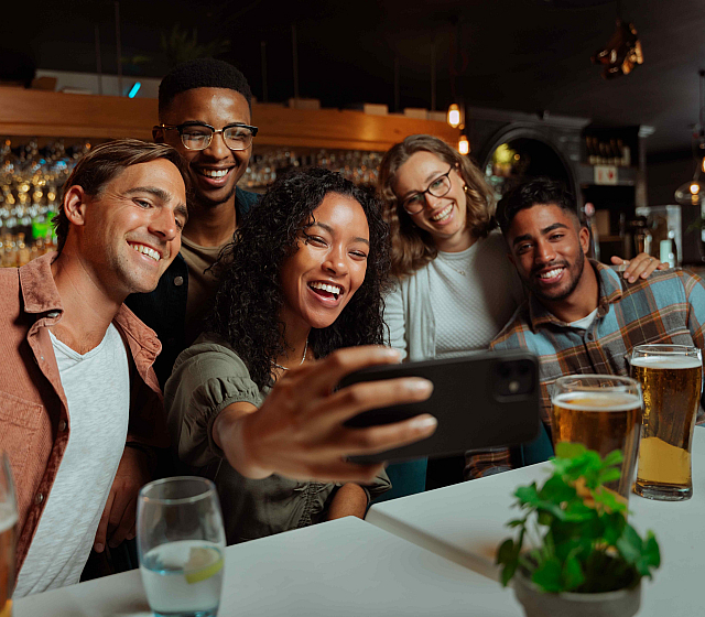 Group of friends smiling and taking a selfie in a lively pub atmosphere, surrounded by glasses of beer and a lively bar background.