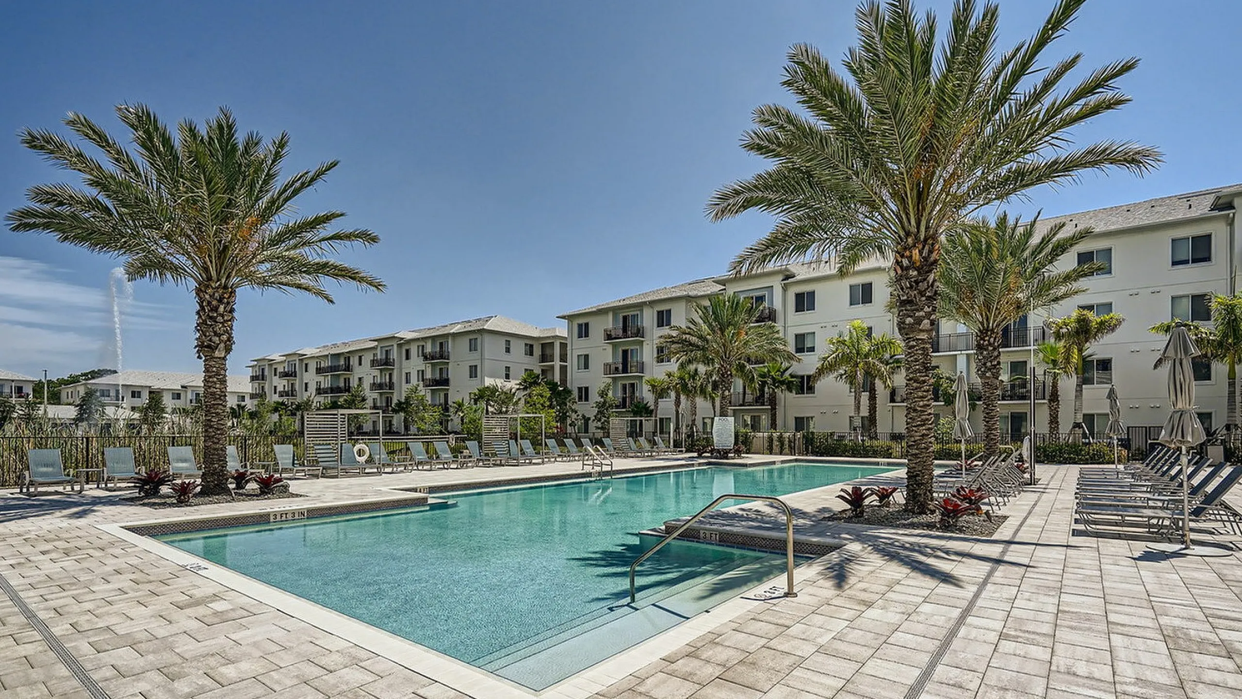 Luxury apartment complex with palm-lined pool, sun loungers, and modern architecture under a clear blue sky.