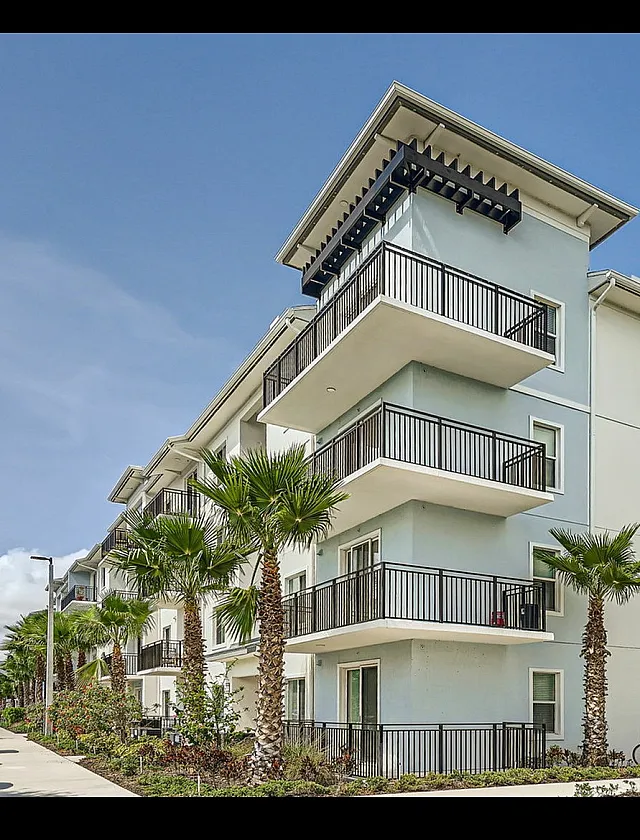 Modern apartment building with spacious balconies, surrounded by lush palm trees and vibrant landscaping under a clear blue sky.