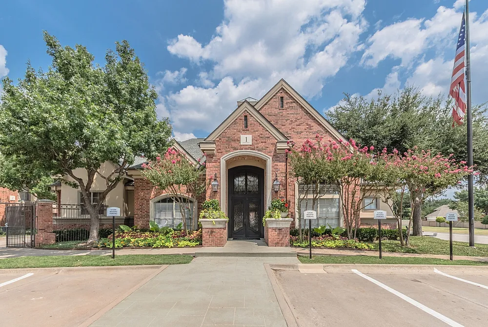 Charming, red-brick apartment facade with lush landscaping, flowering trees, and vibrant curb appeal under a bright blue sky.