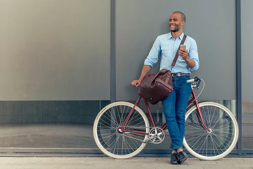 Confident man in a light blue shirt holding a phone, casually leaning against a vintage bicycle and smiling.