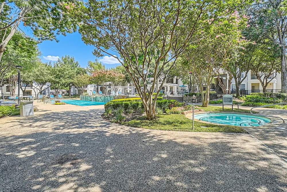 Inviting apartment complex with lush greenery, a sparkling swimming pool, and serene walkways under a clear blue sky.