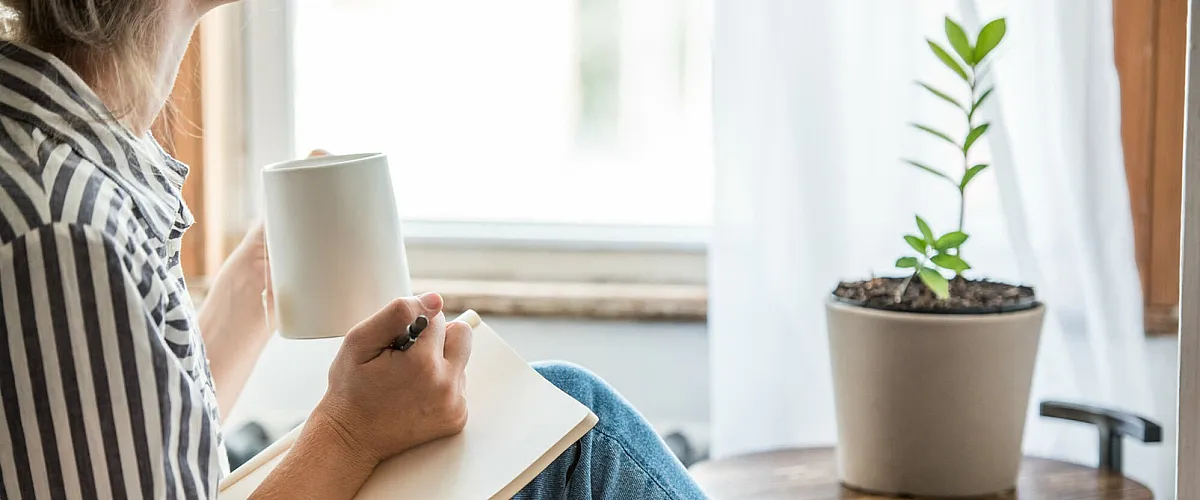 Cozy apartment scene with a person enjoying a warm drink and writing, next to a vibrant potted plant on a stylish wooden table.
