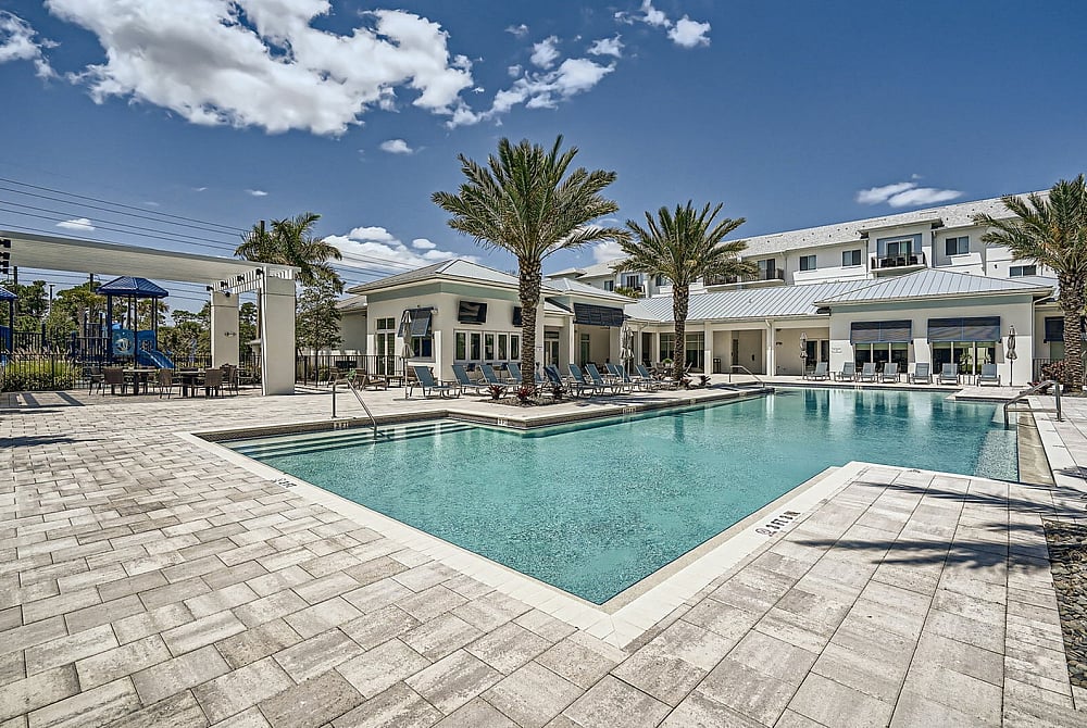 Luxurious apartment complex pool area with palm trees, lounge chairs, and modern architecture under a clear blue sky.