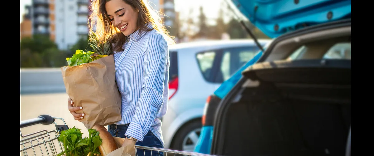 Cheerful woman in sunny parking lot loading groceries into her car, highlighting a convenient lifestyle near urban apartments.