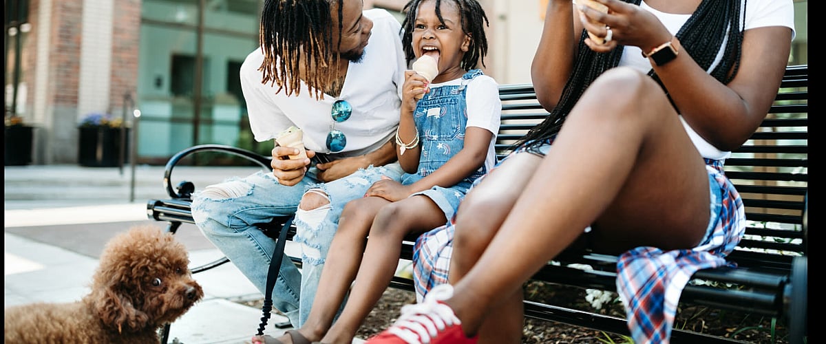 Joyful family moment as a father, mother, and daughter enjoy ice cream together on a sunny day, accompanied by their cute poodle, outside a modern building.