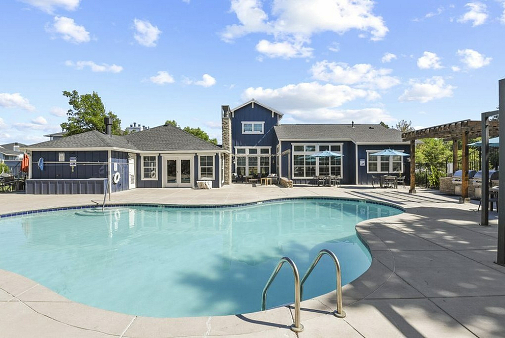 Charming outdoor view of a luxurious apartment complex featuring a sparkling swimming pool, inviting lounge area, and stylish architecture under a bright blue sky.