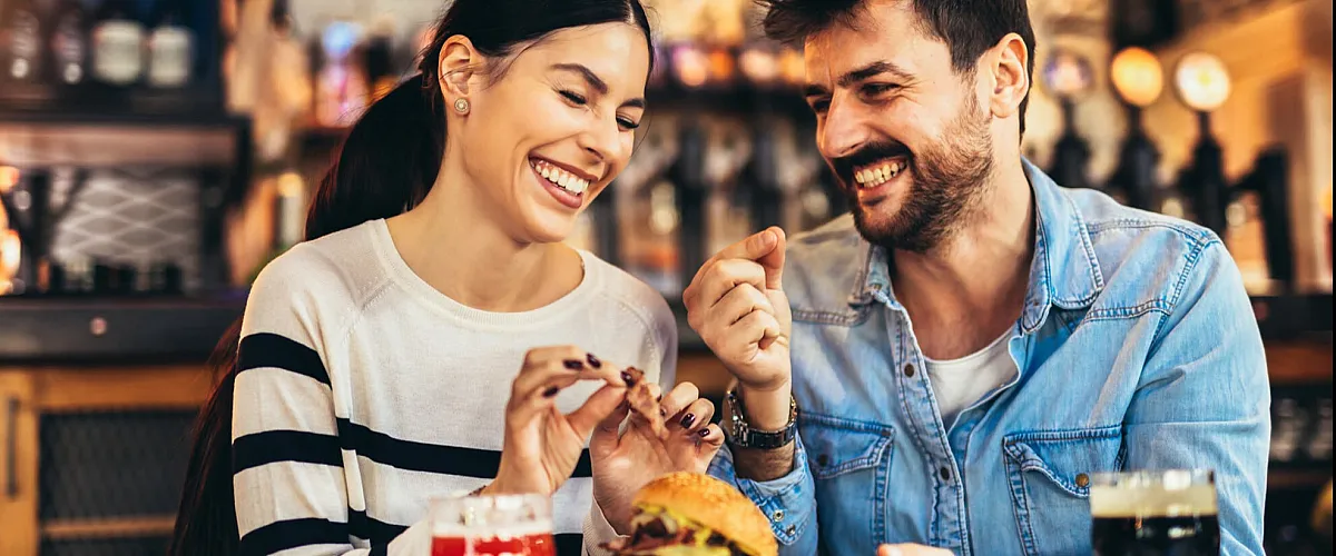 Cheerful couple enjoying delicious burgers and drinks at a lively bar setting.