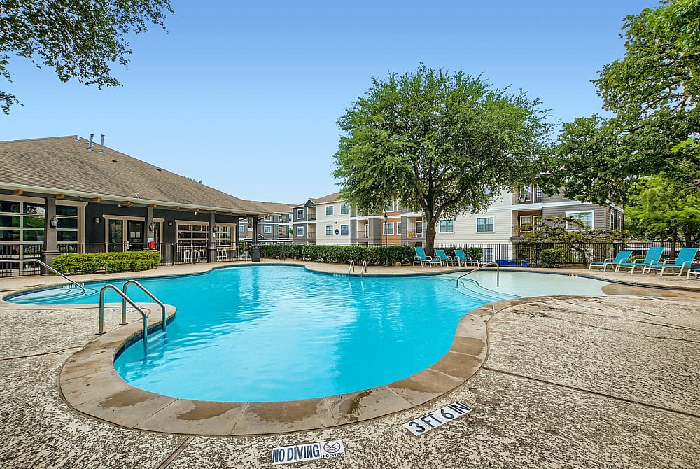 Inviting apartment complex pool with sun loungers, surrounded by lush greenery and modern buildings, perfect for relaxation.