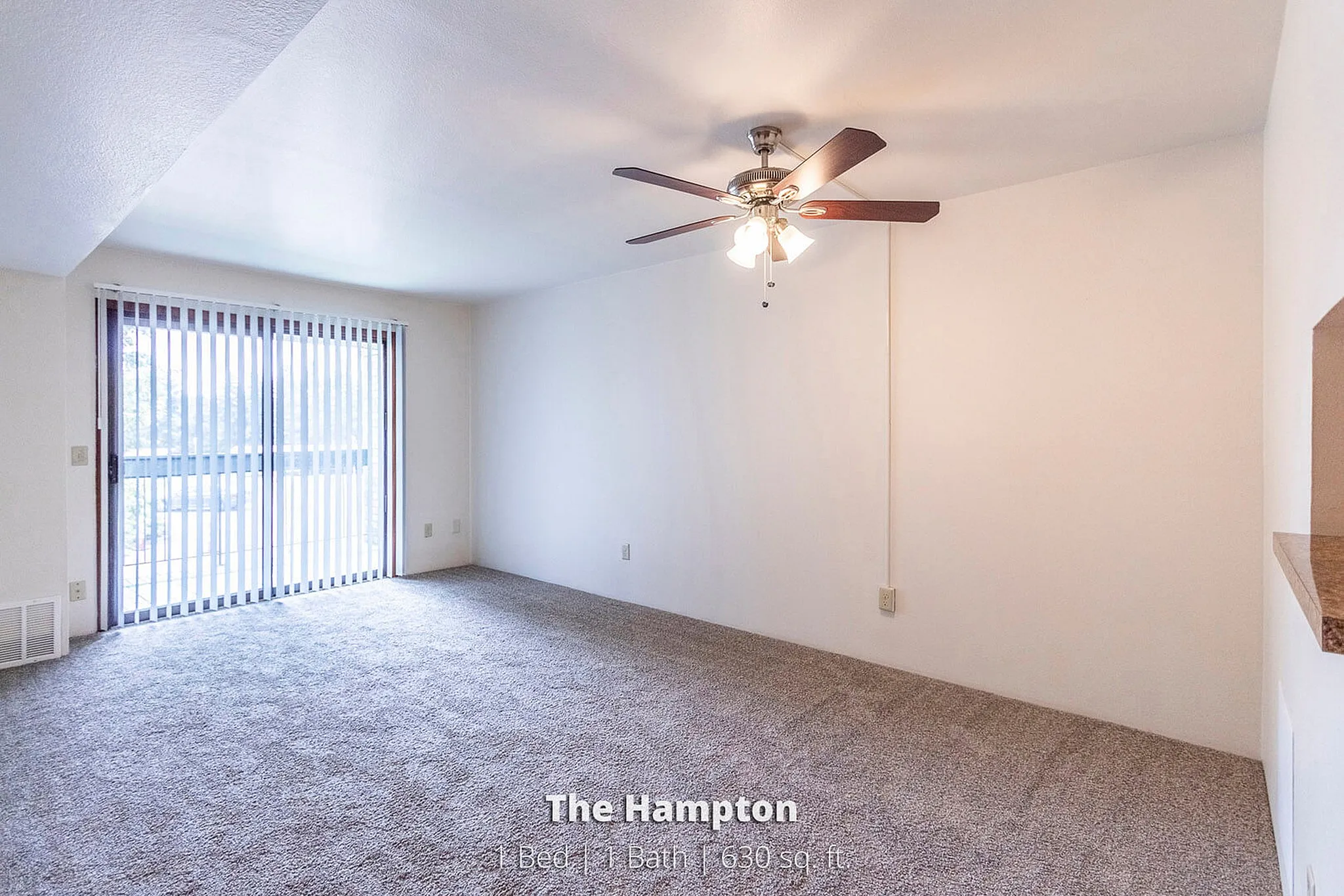 Bright and inviting living room in The Hampton apartment, featuring plush carpeting and a ceiling fan, ideal for comfort. Large window with vertical blinds offers abundant natural light in this spacious 1 bed, 1 bath layout.