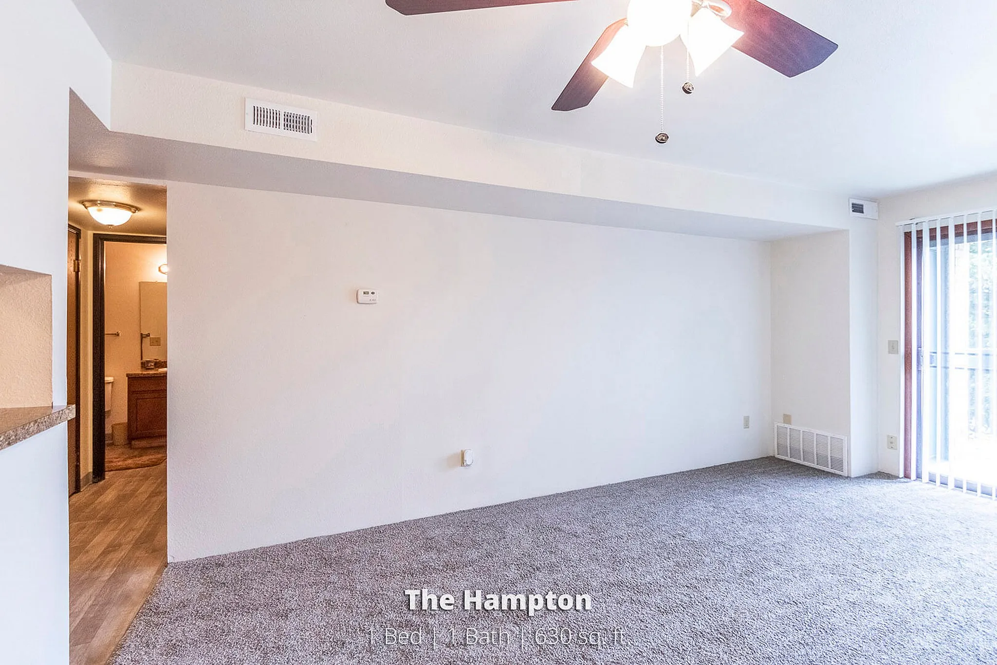 Bright and welcoming living space in The Hampton, featuring modern ceiling fan, plush carpet, and ample natural light from large windows. 1 bed, 1 bath, 630 sq. ft.