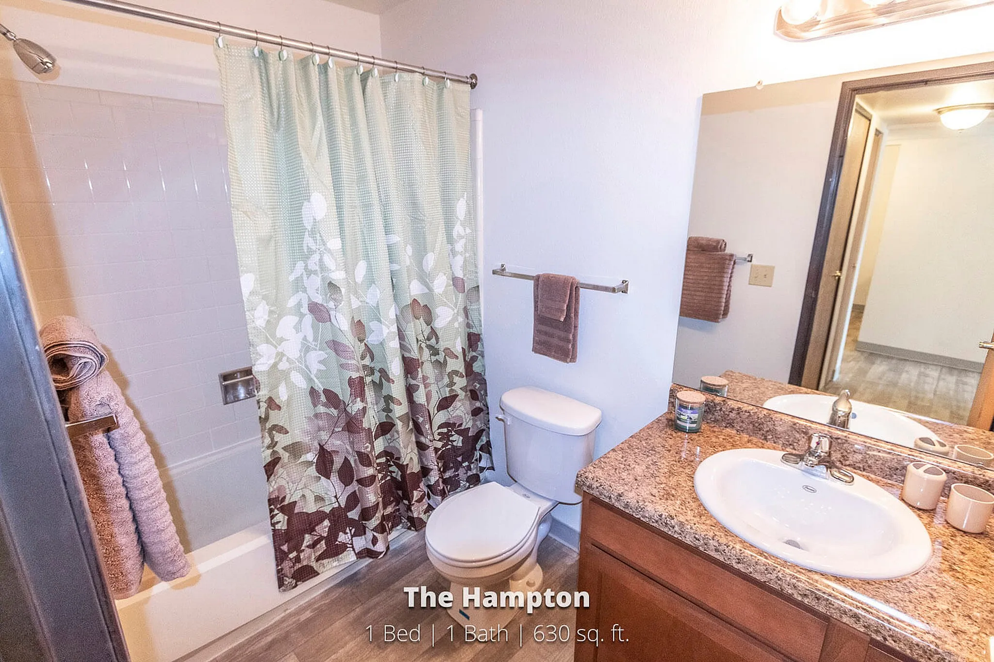 Bright and modern bathroom in 'The Hampton' 1 Bed, 1 Bath apartment, featuring elegant granite countertops, stylish floral shower curtain, and cozy lighting over a spacious mirror.