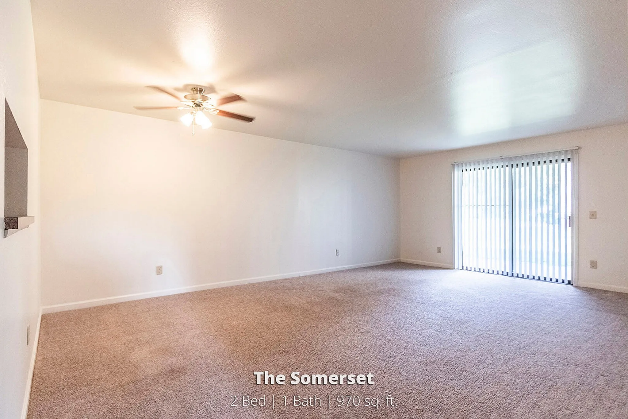 Bright and spacious living room in The Somerset with carpeted floors, ceiling fan, and expansive glass doors leading to an outdoor view. Ideal for modern living in 970 sq. ft.