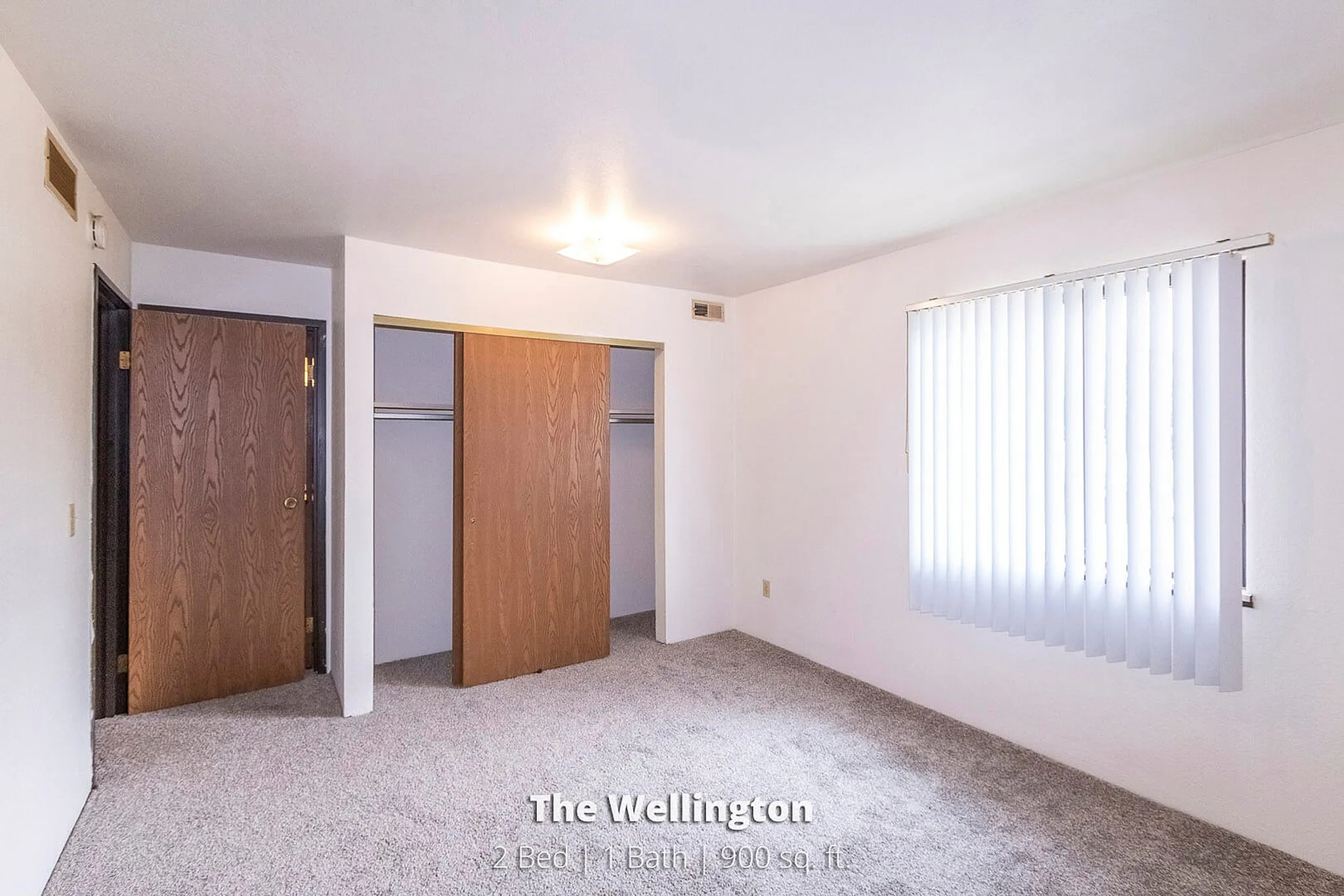 Bright and spacious bedroom in The Wellington, featuring a large window with vertical blinds, ample closet space, and cozy carpeting. Perfect for comfort and relaxation in this 2 bed, 1 bath, 900 sq. ft. home.