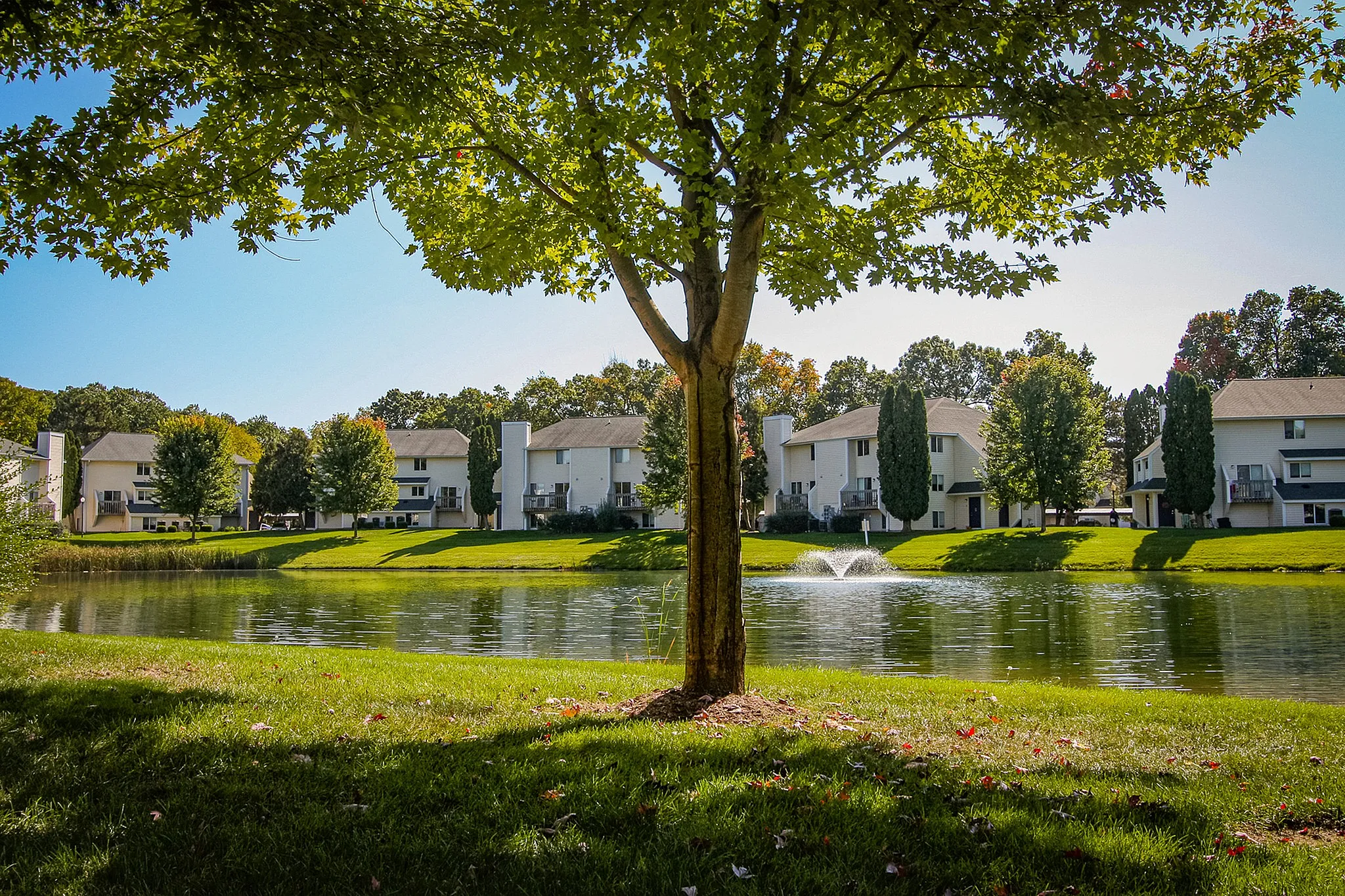 Charming lakeside apartment community with lush greenery, fountain, and vibrant trees under a clear blue sky.