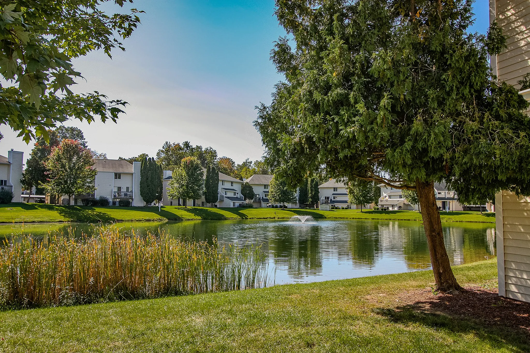 Scenic view of tranquil lakeside apartments surrounded by lush greenery and a serene pond with a fountain.