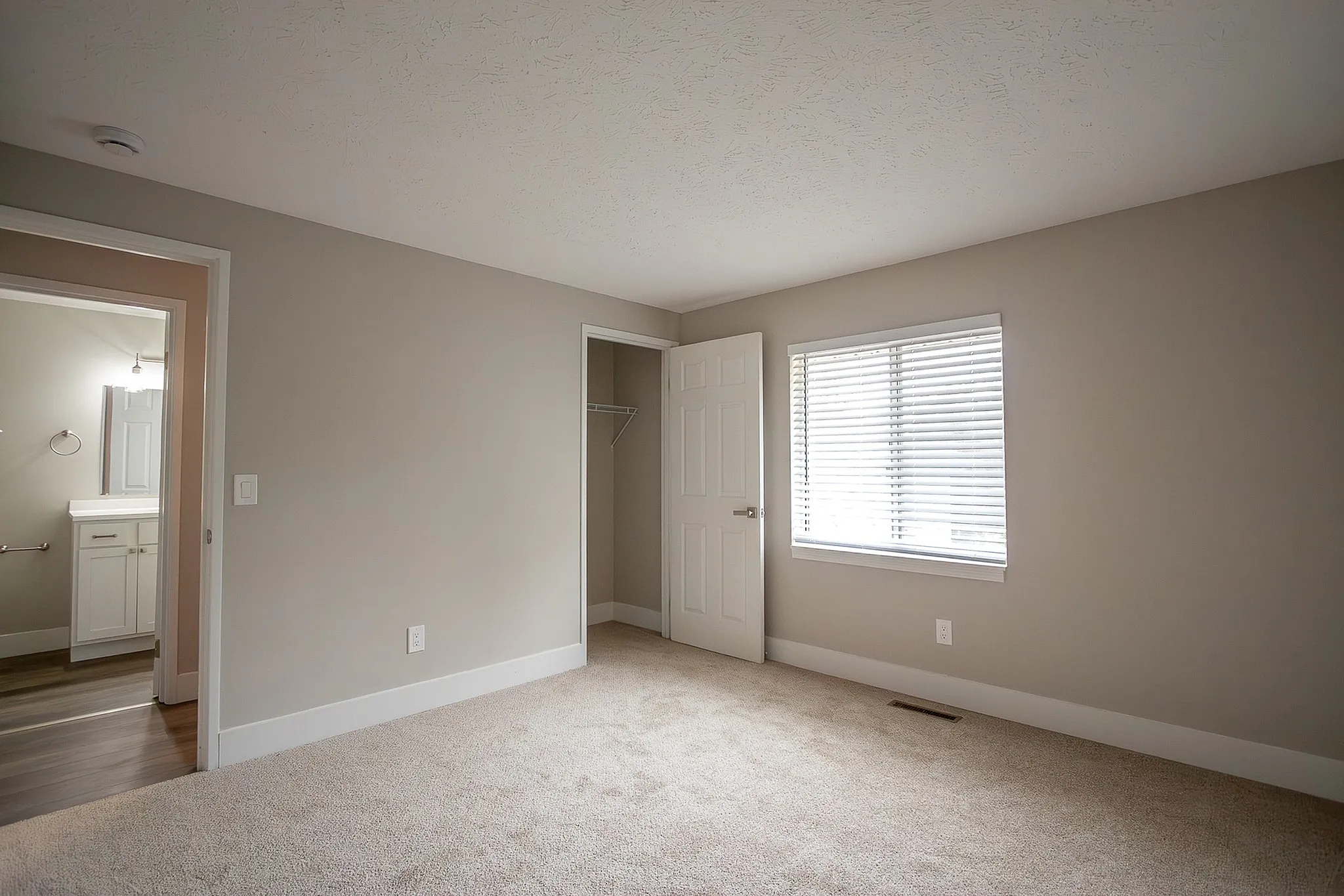 Bright and inviting bedroom featuring plush carpeting, large window with blinds, and an open closet, perfect for modern living.