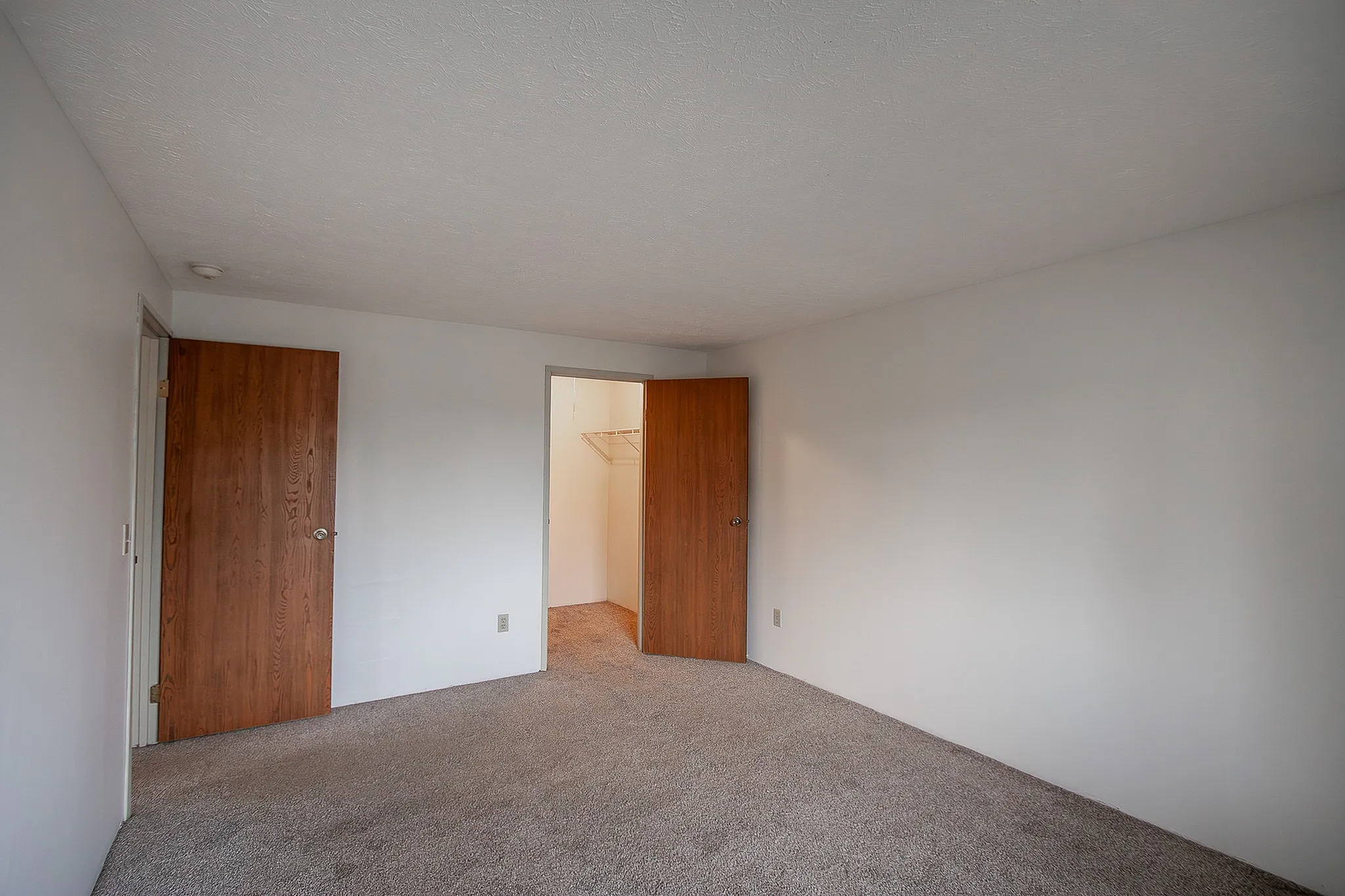 Spacious bedroom with warm wooden doors leading to closets, featuring soft carpet and a bright, neutral ambiance.