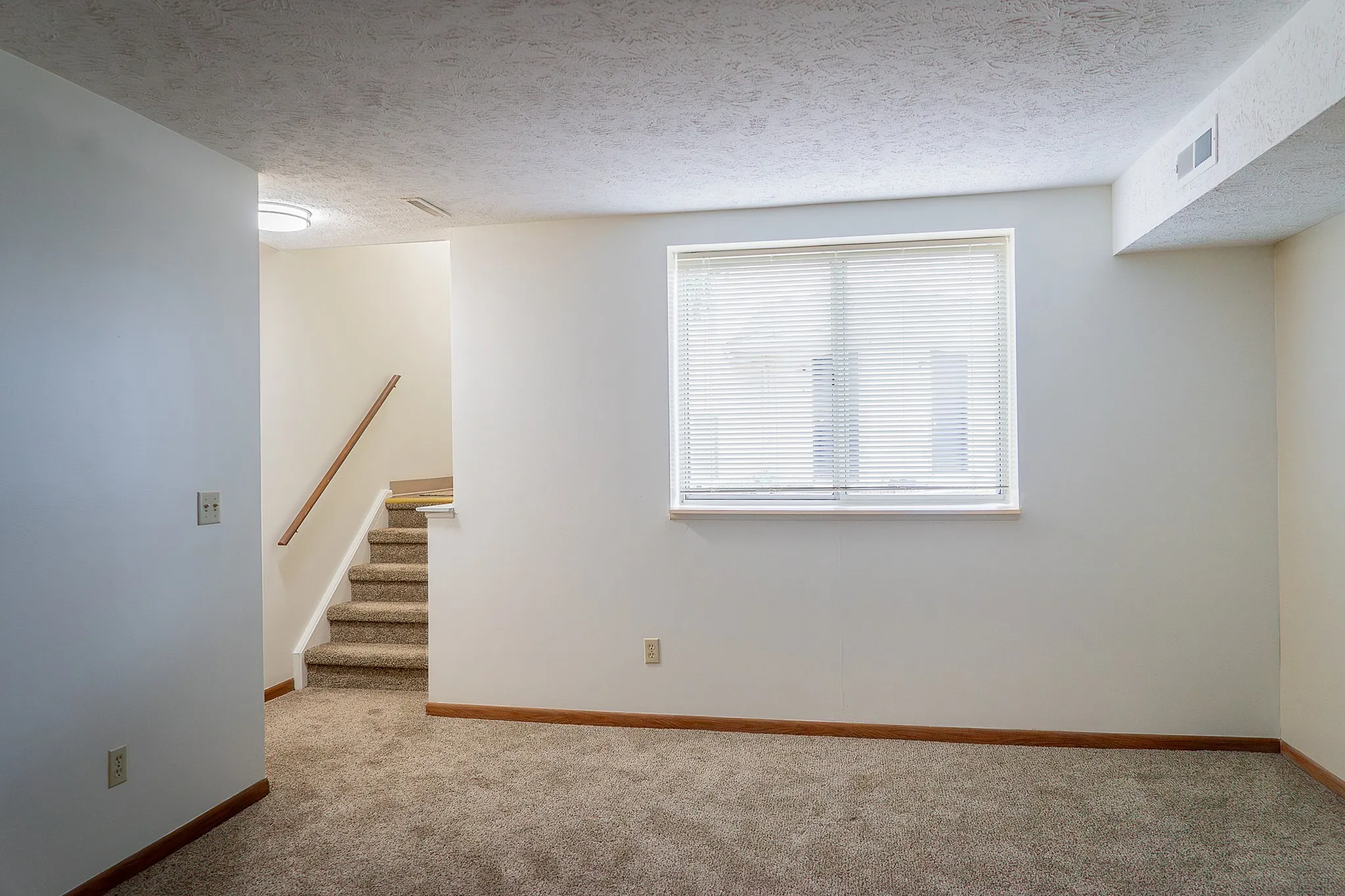 Bright, cozy room with carpet flooring, large window with blinds, and a carpeted staircase leading upward.