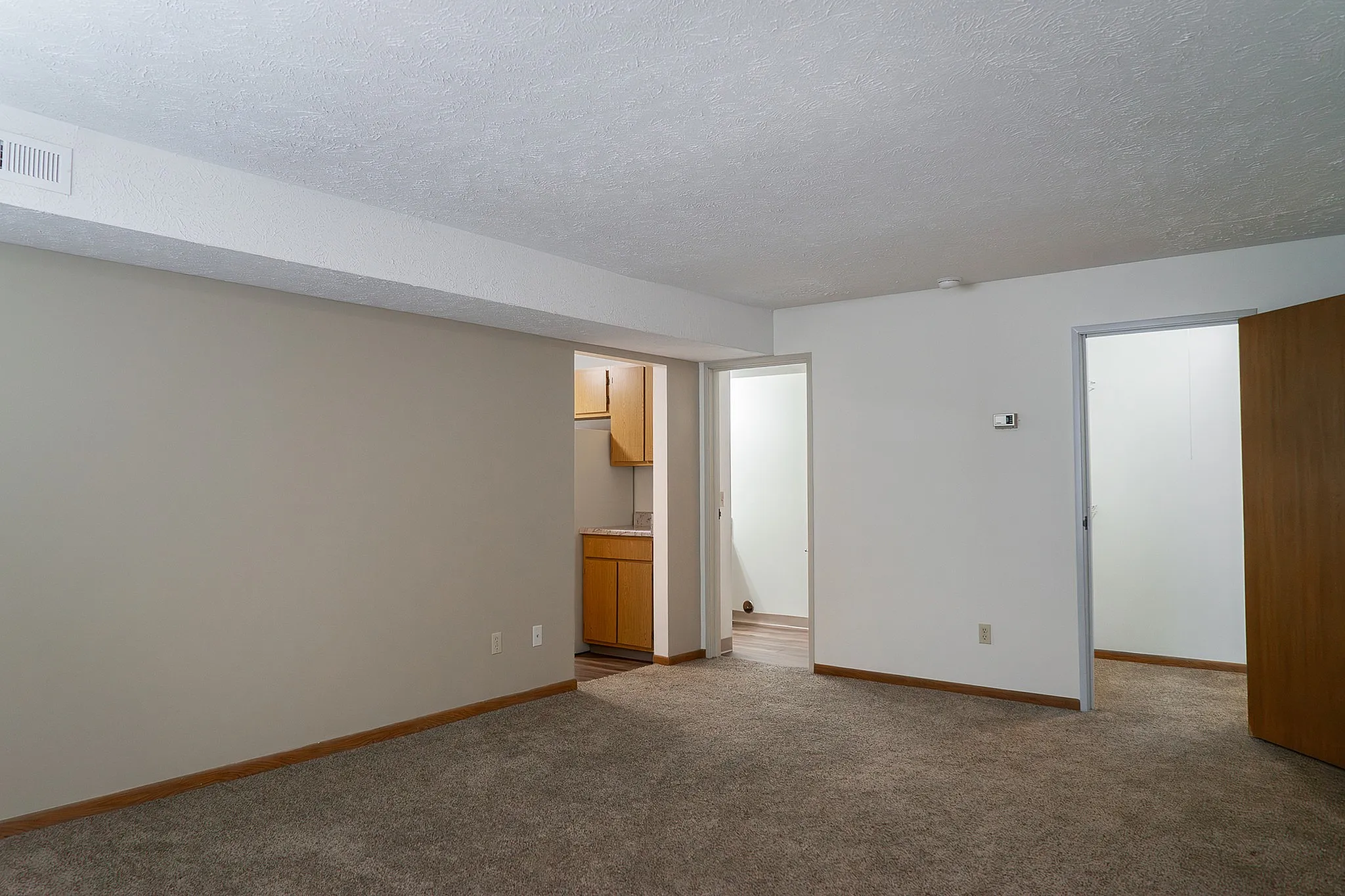 Inviting apartment with plush carpeted floors, warm wall tones, and an open doorway leading to a cozy kitchen, perfect for comfortable living.