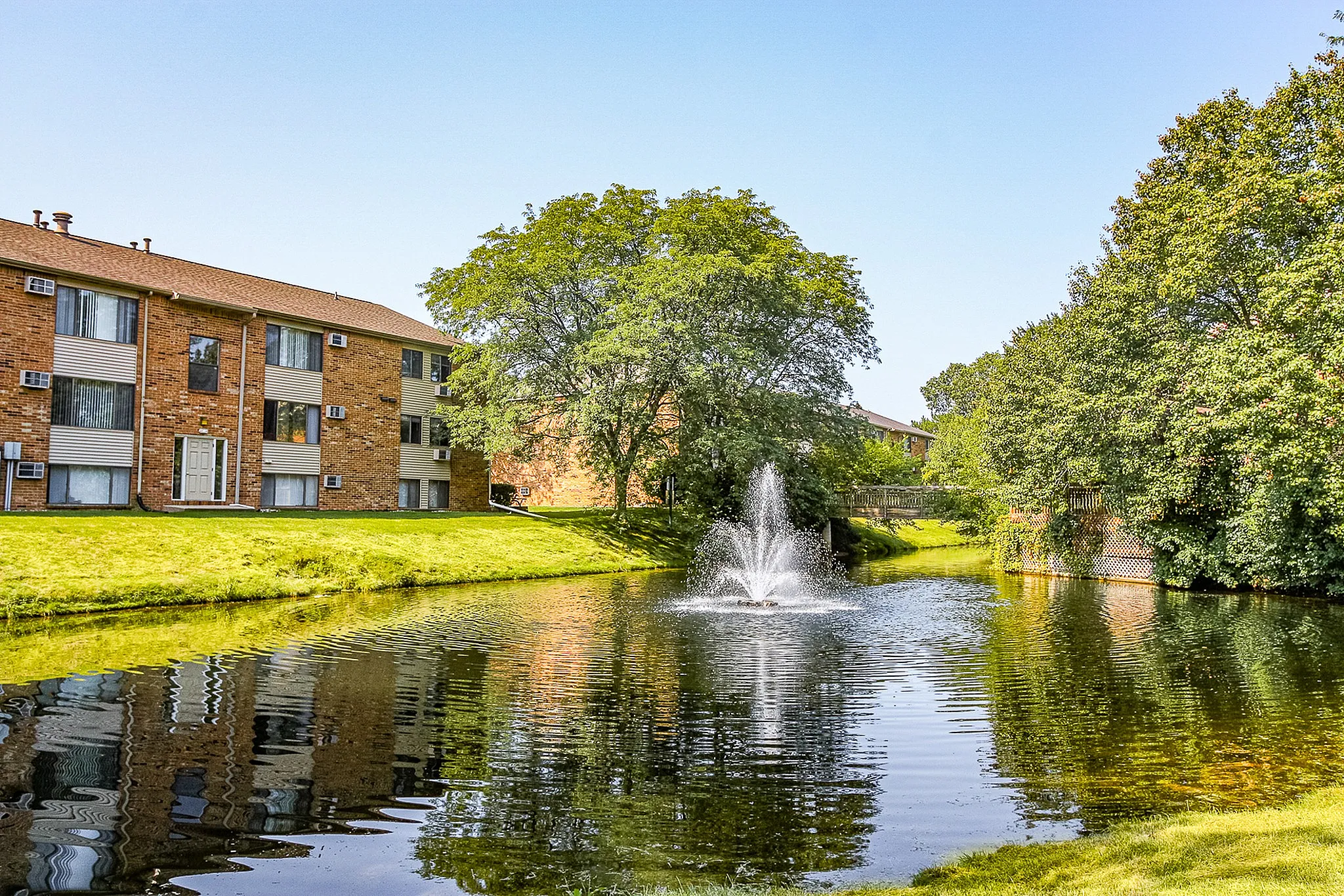 Charming apartment complex nestled by a tranquil pond with a stunning fountain, surrounded by lush greenery.