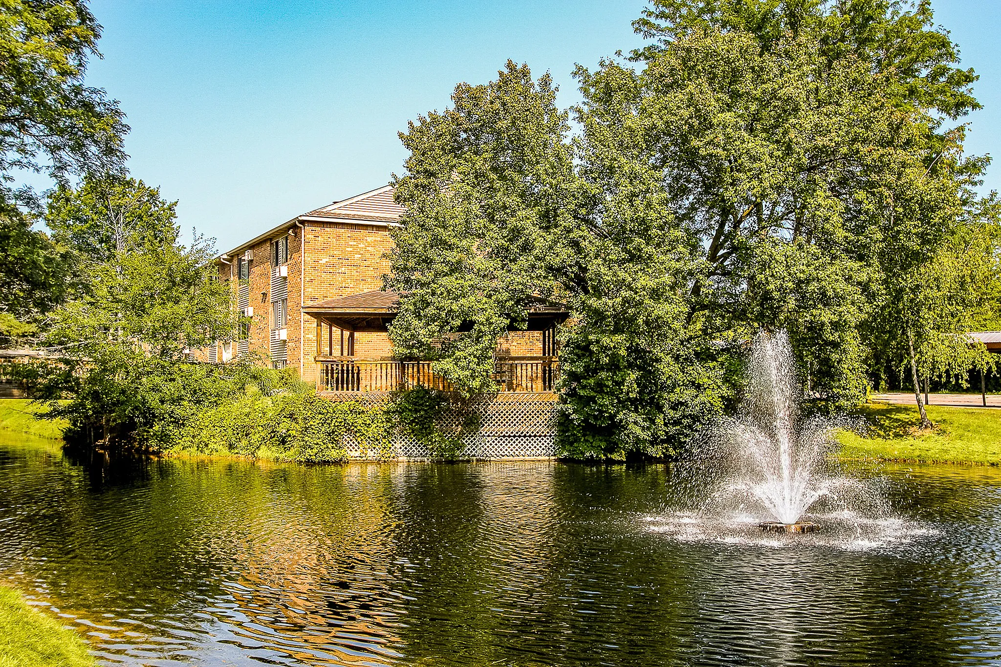 Charming brick apartment building with a private balcony overlooking a serene pond and fountain, surrounded by lush greenery.