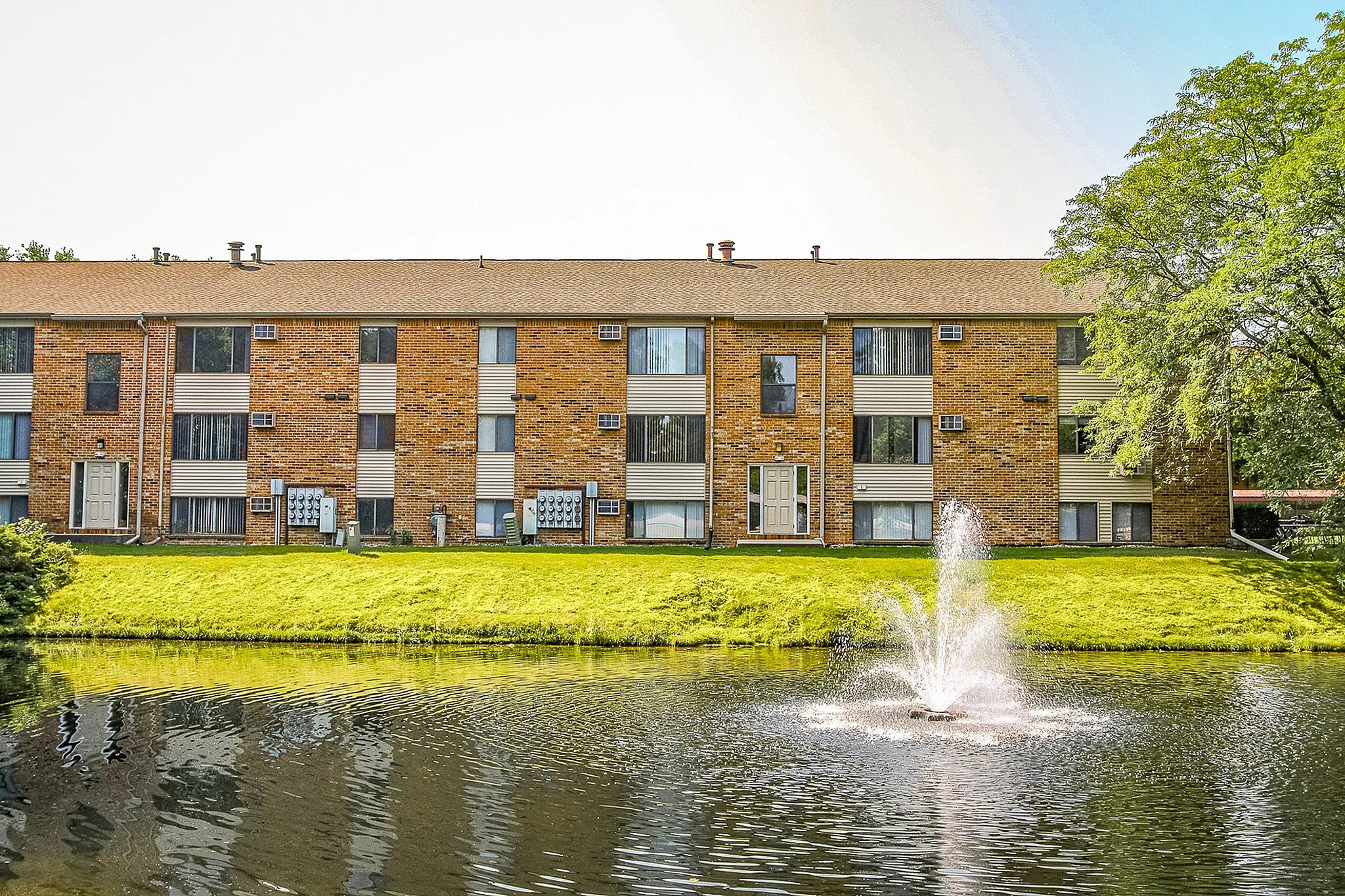 Charming brick apartment complex featuring serene pond and a beautiful fountain in a lush green setting.