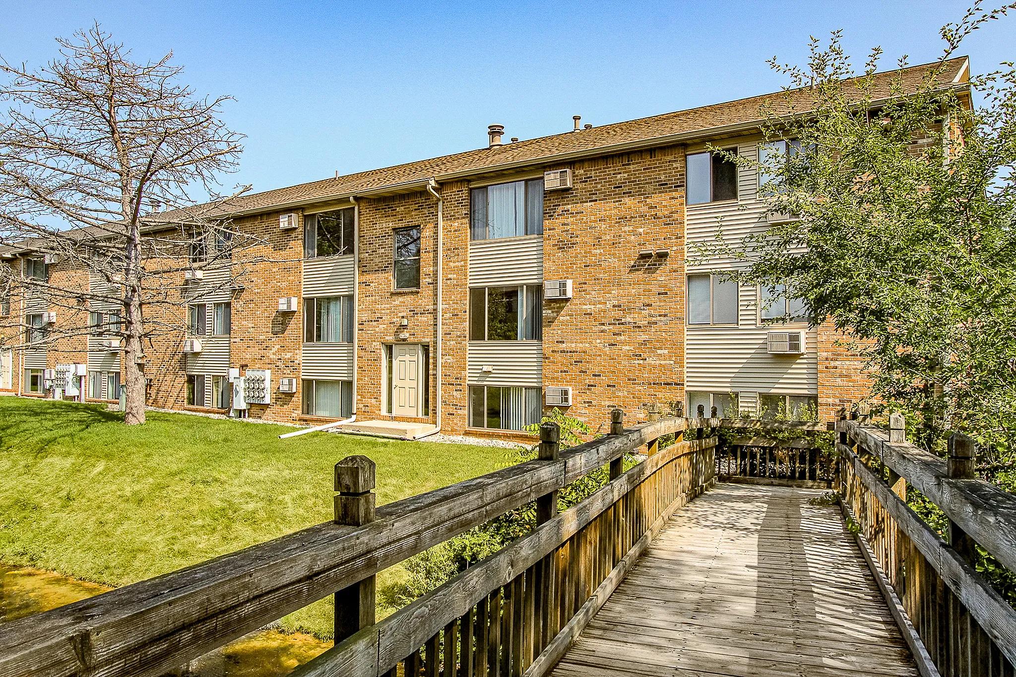 Inviting brick apartment complex with lush green lawns and serene wooden walkway, exuding charm and comfort.