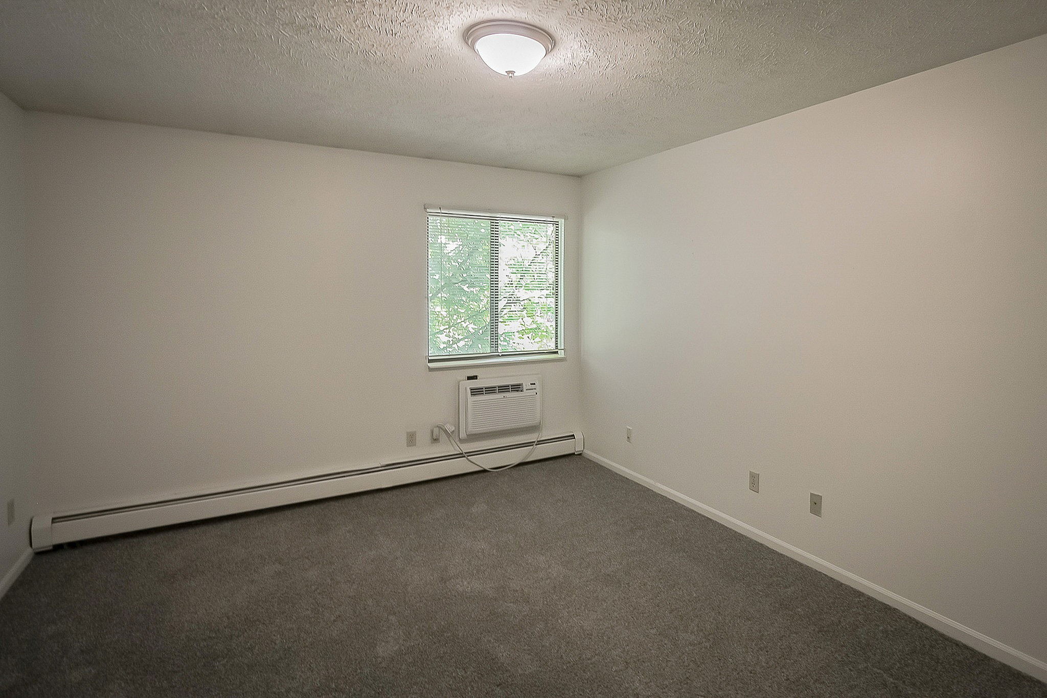 Bright and airy room with soft carpeting, a large window for natural light, and a built-in air conditioning unit, perfect for creating a cozy retreat.