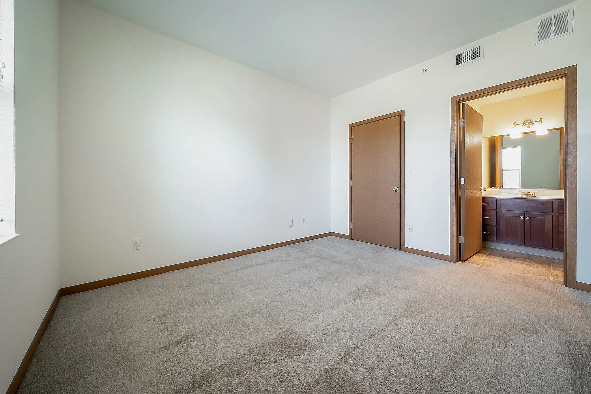 Bright, spacious bedroom with plush carpeting, featuring an en-suite bathroom with warm wooden cabinetry.