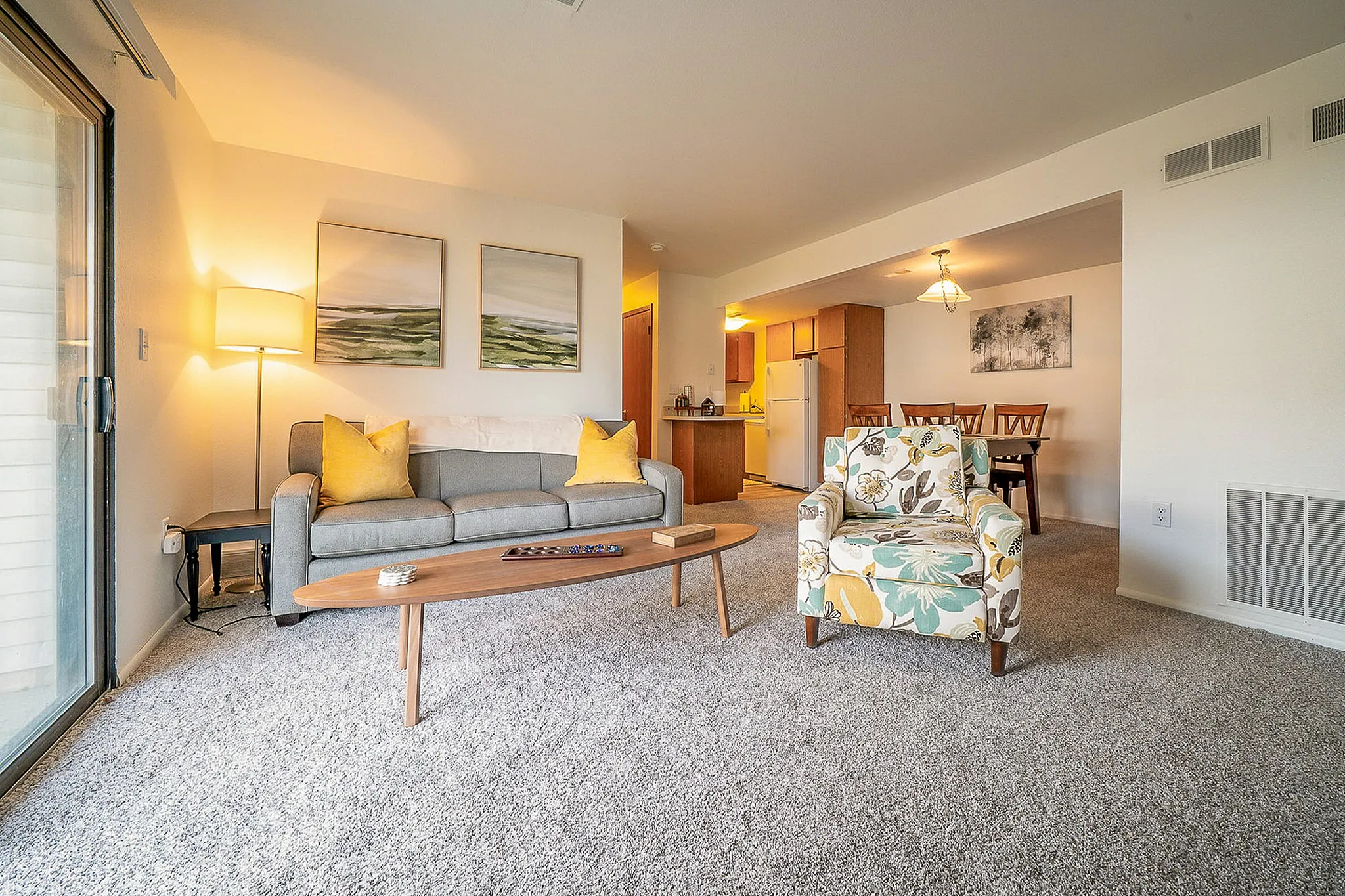 Bright and inviting living room with plush carpeting, featuring a modern grey sofa with yellow cushions, a floral armchair, and a sleek wooden coffee table. The open-concept layout elegantly extends to a cozy dining area and kitchen, enhanced by ambient lighting and contemporary artwork.