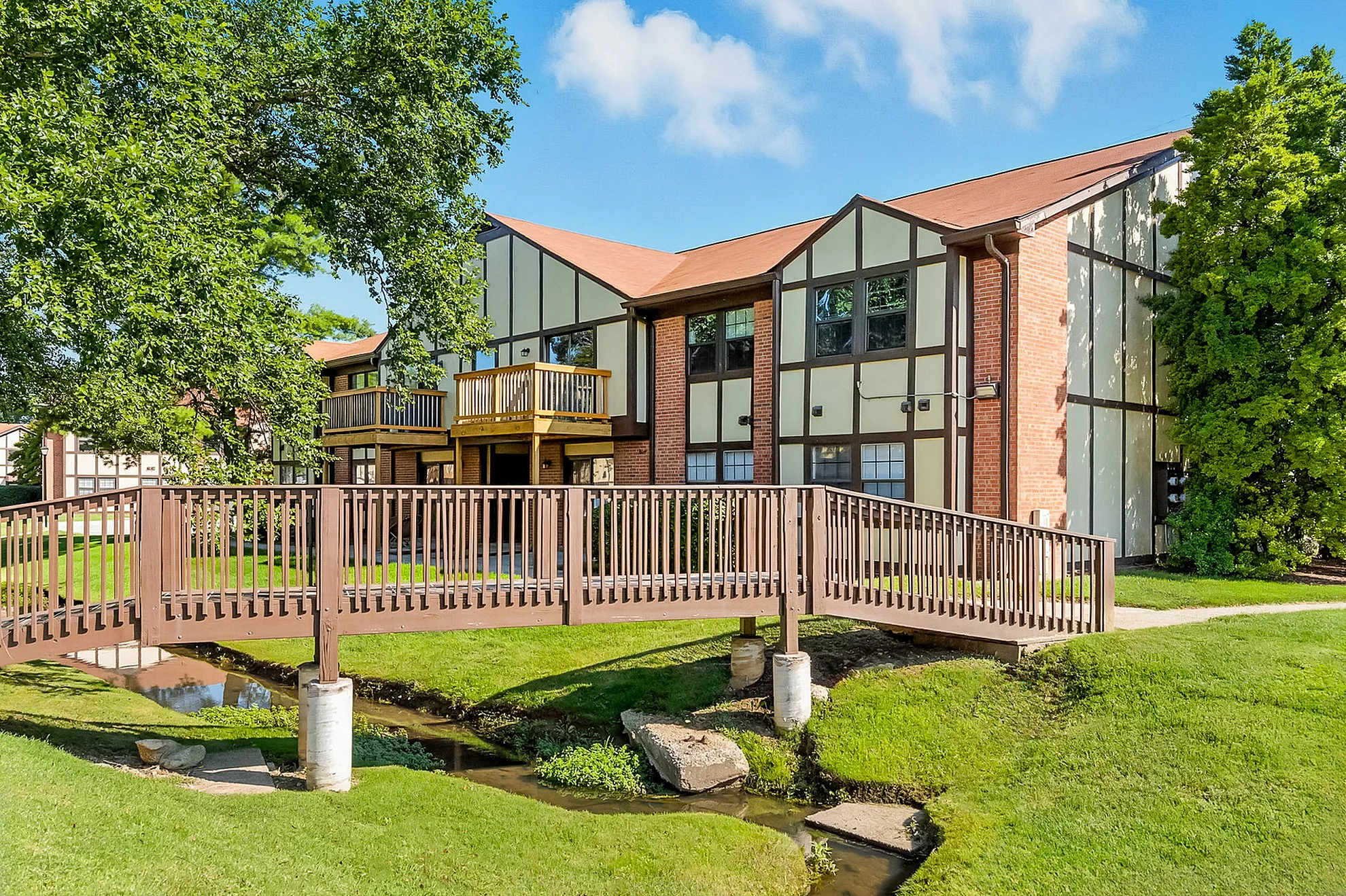 Charming apartment complex featuring classic Tudor architecture, lush green lawns, and a picturesque wooden bridge.